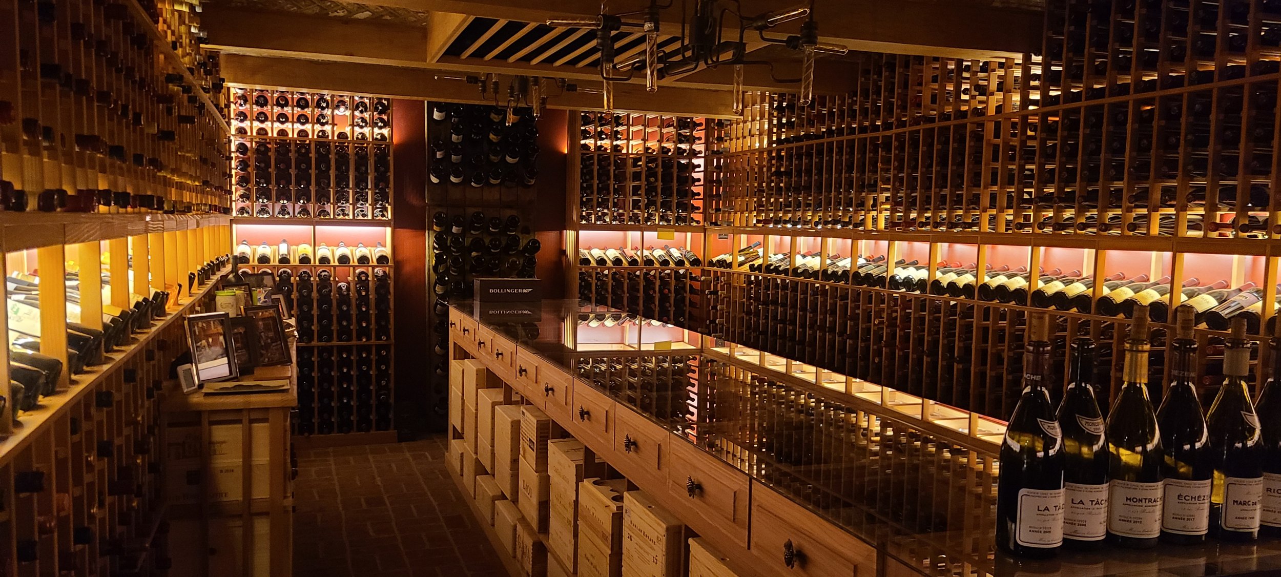 A wine cellar with wooden racks filled with bottles of wine, illuminated by soft lighting. Bottles are stored horizontally, with some displayed upright on a counter.