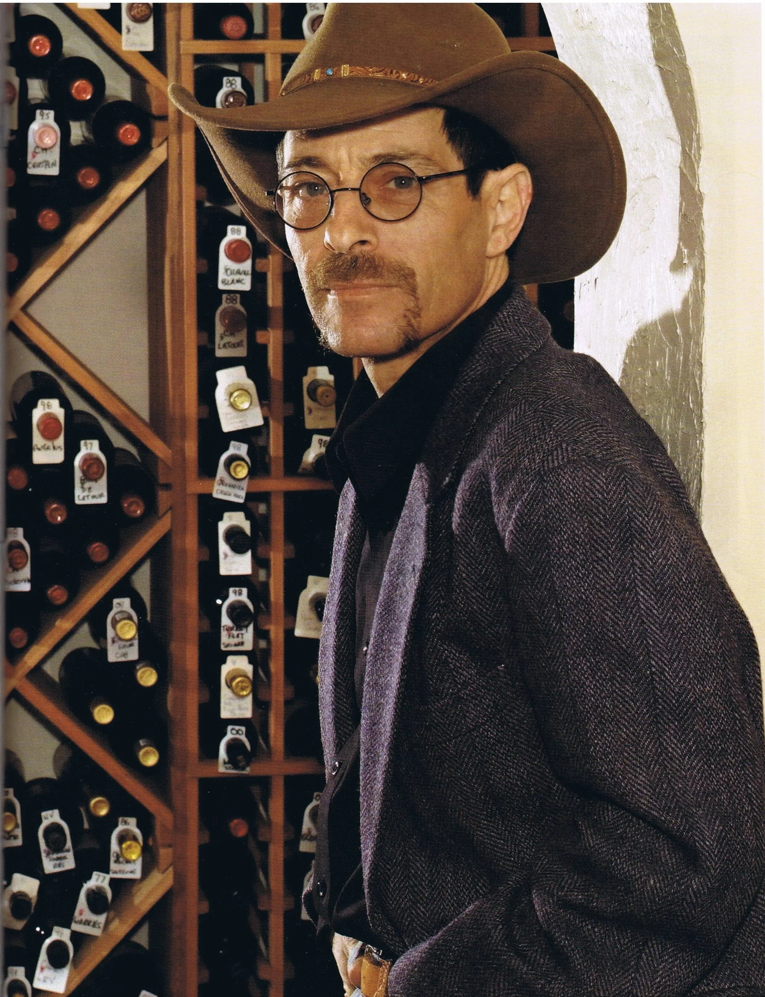 A man wearing a cowboy hat, glasses, a dark shirt, and a blazer standing in front of a wine rack.