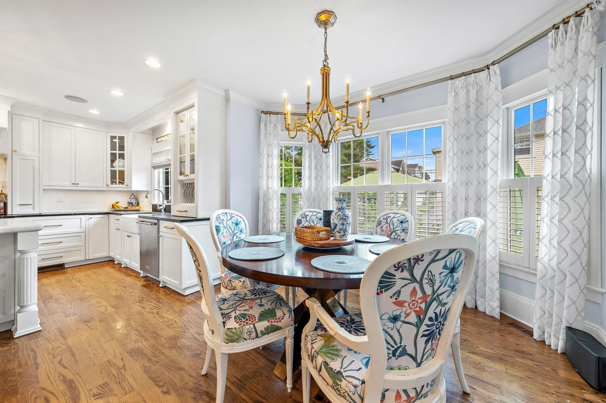 Bright dining area with large windows, white curtains, hardwood floor, round wooden table with six floral-patterned chairs, gold chandelier hanging above, and a white kitchen in the background.