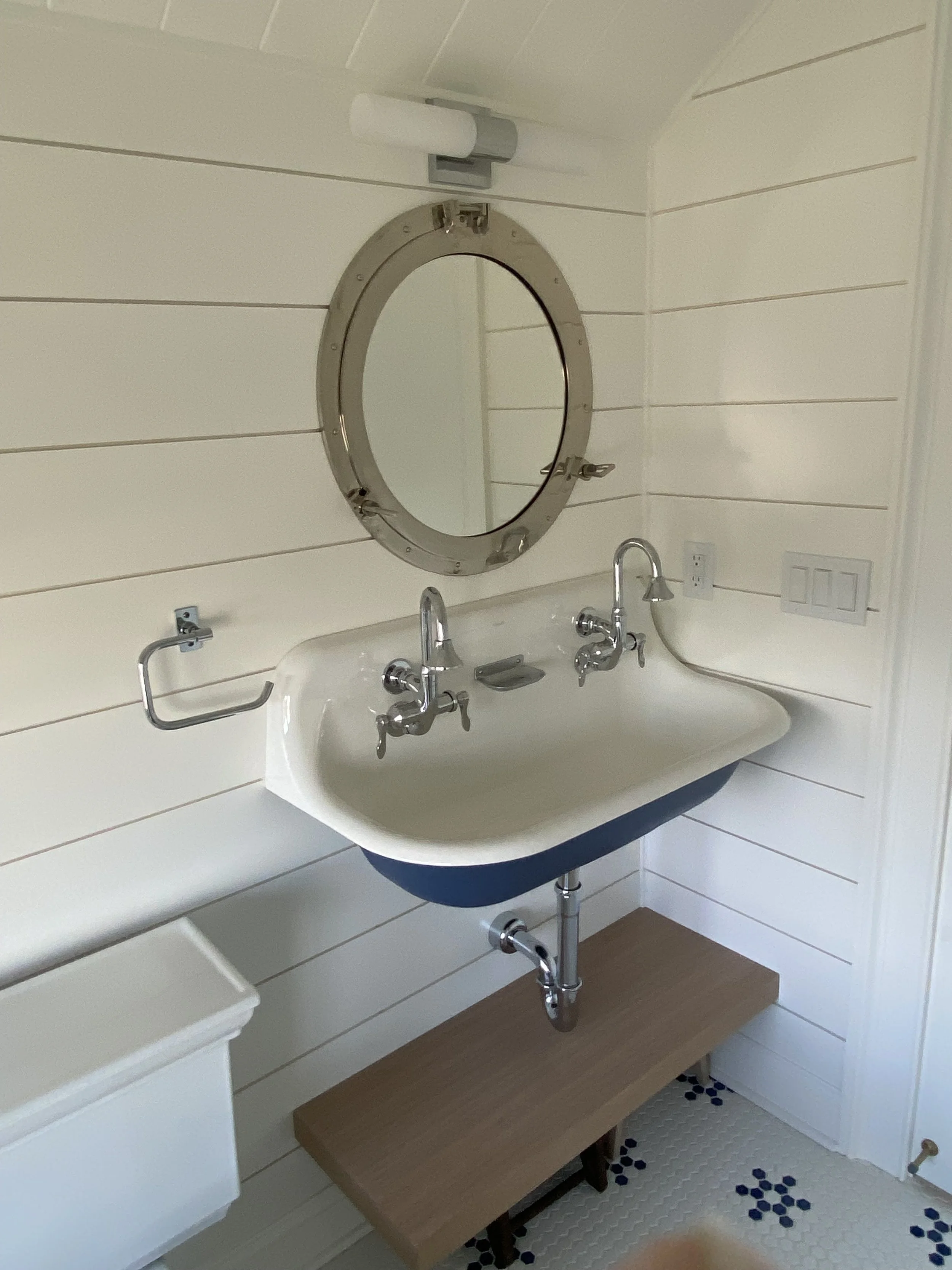 A vintage bathroom sink with two separate faucets, a mirror with a nautical detail, and a wooden shelf below. The walls are done in ship-lap which portrays a clean coastal esthetic. 