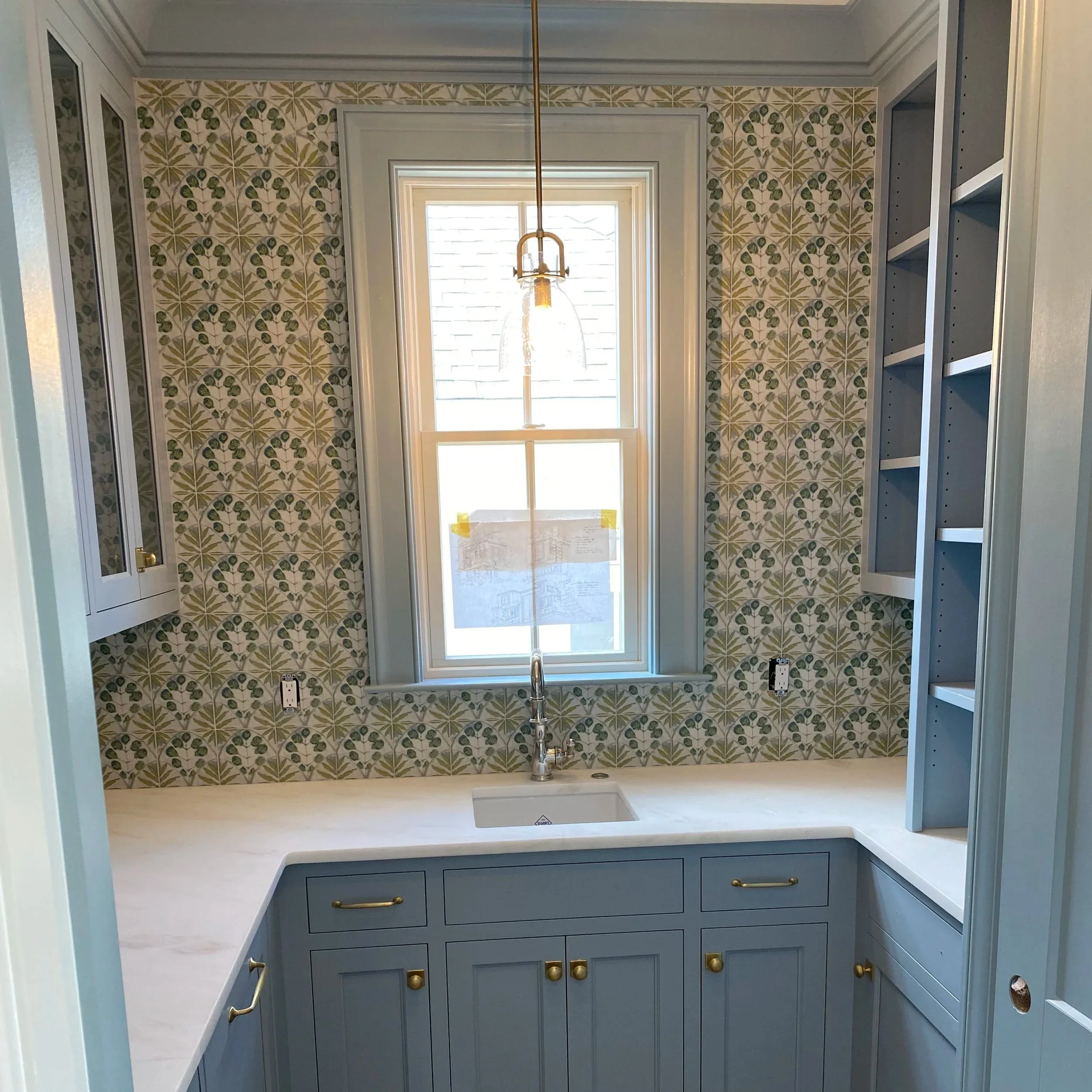 A kitchen with a large window above the sink, a hanging pendant light, blue cabinets, and a patterned wallpaper with green and beige leaves.