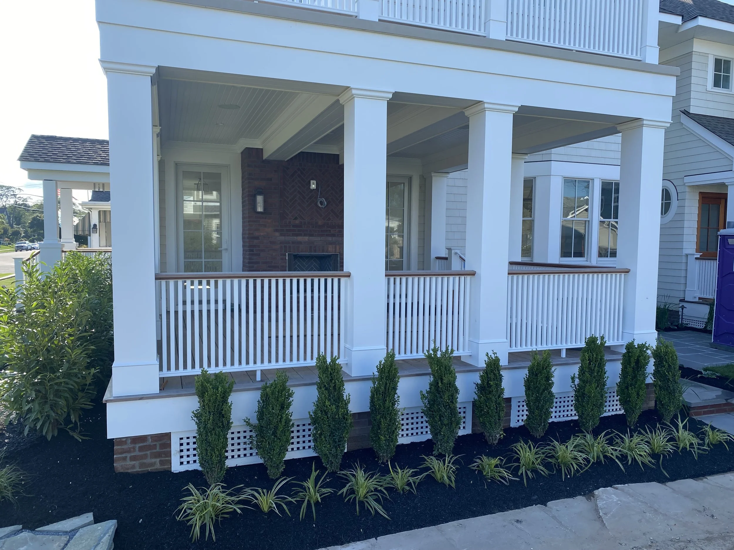 Front porch of a modern house with white railing, brick chimney, and surrounding landscaped garden with small shrubs and plants.