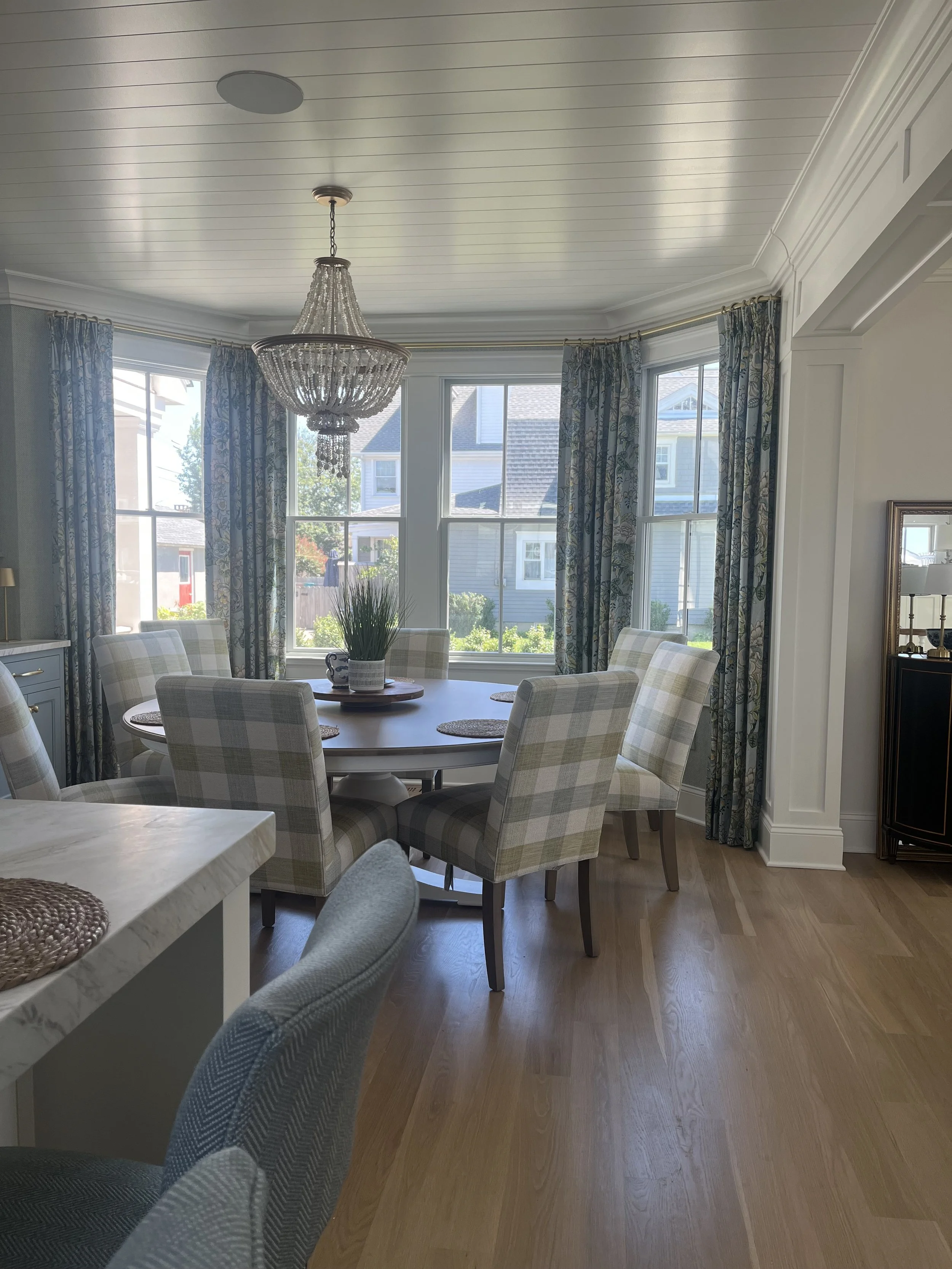 Bright dining room with a round table and six plaid upholstered chairs, large bay window with drapes, coastal inspired chandelier and hardwood flooring.