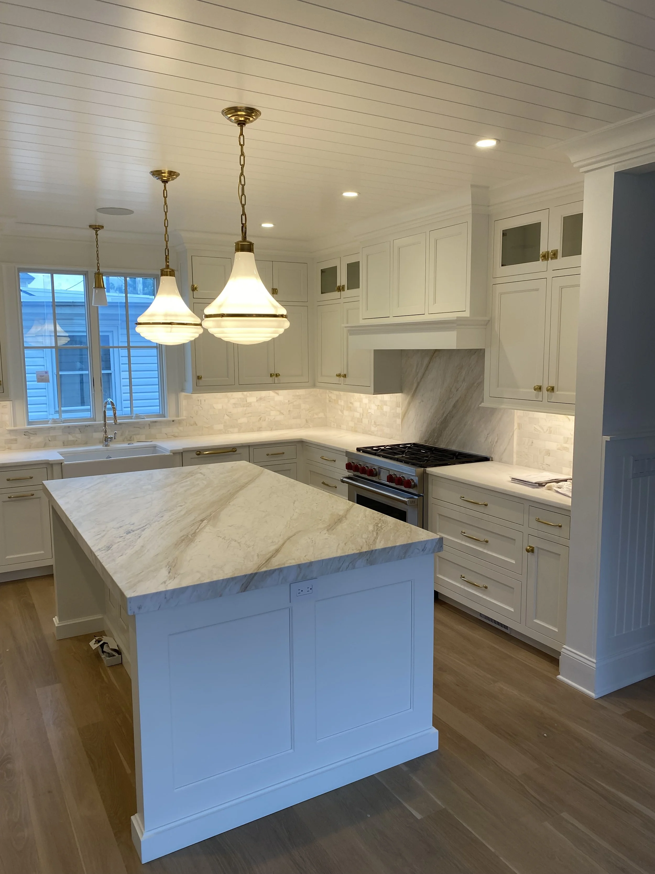 Bright white kitchen with marble countertops and a central island, gold hardware, pendant lights, and wood flooring.