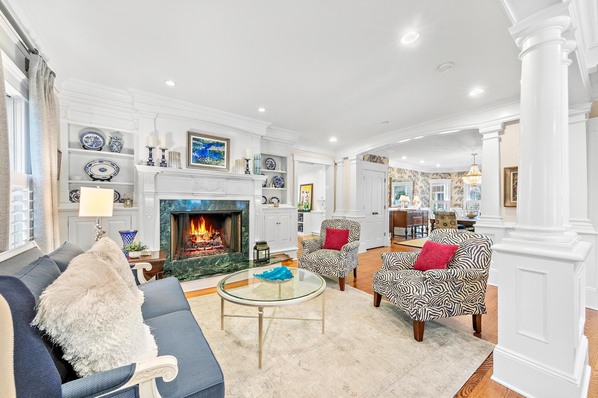 Bright living room with white walls, a fireplace with a fire, a blue sofa with a fluffy pillow, and two patterned chairs with red pillows, a glass coffee table, and bookshelves with decorative items.