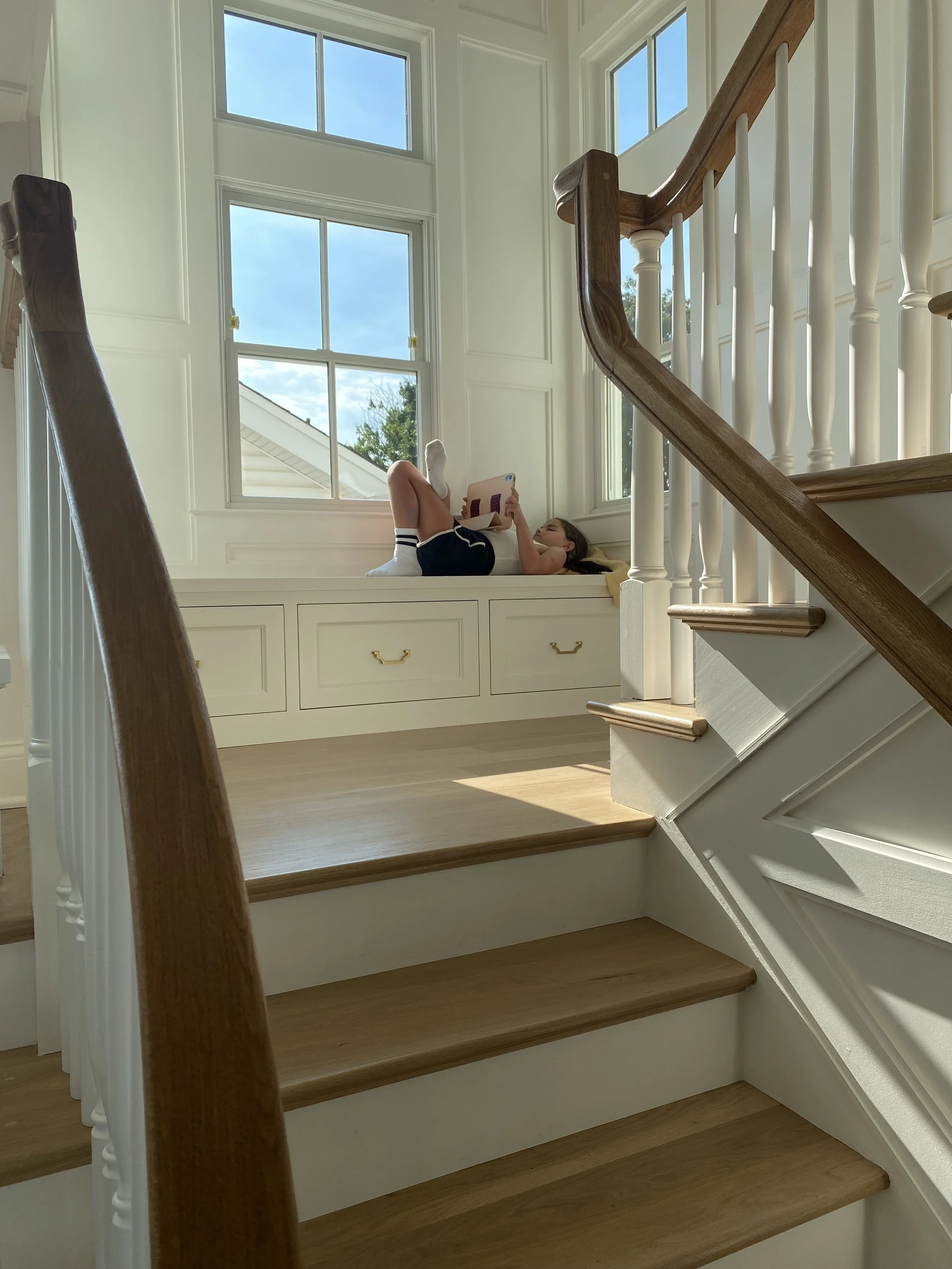 A girl lying on a window seat reading a book in a bright room with large windows and a staircase with wooden steps and white risers.