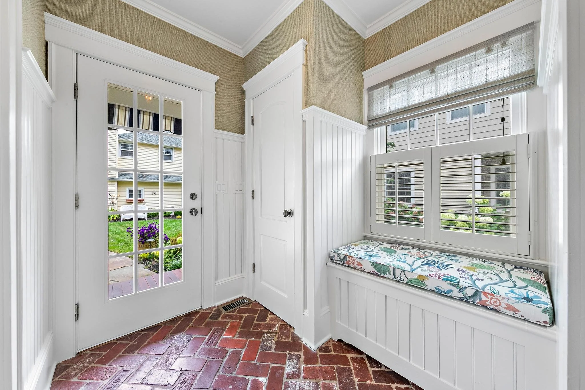 Mud Hall  with brick floor, white bead-board paneling, a french door leading outside, a built-in window seat with colorful floral cushion, and shuttered
window.
