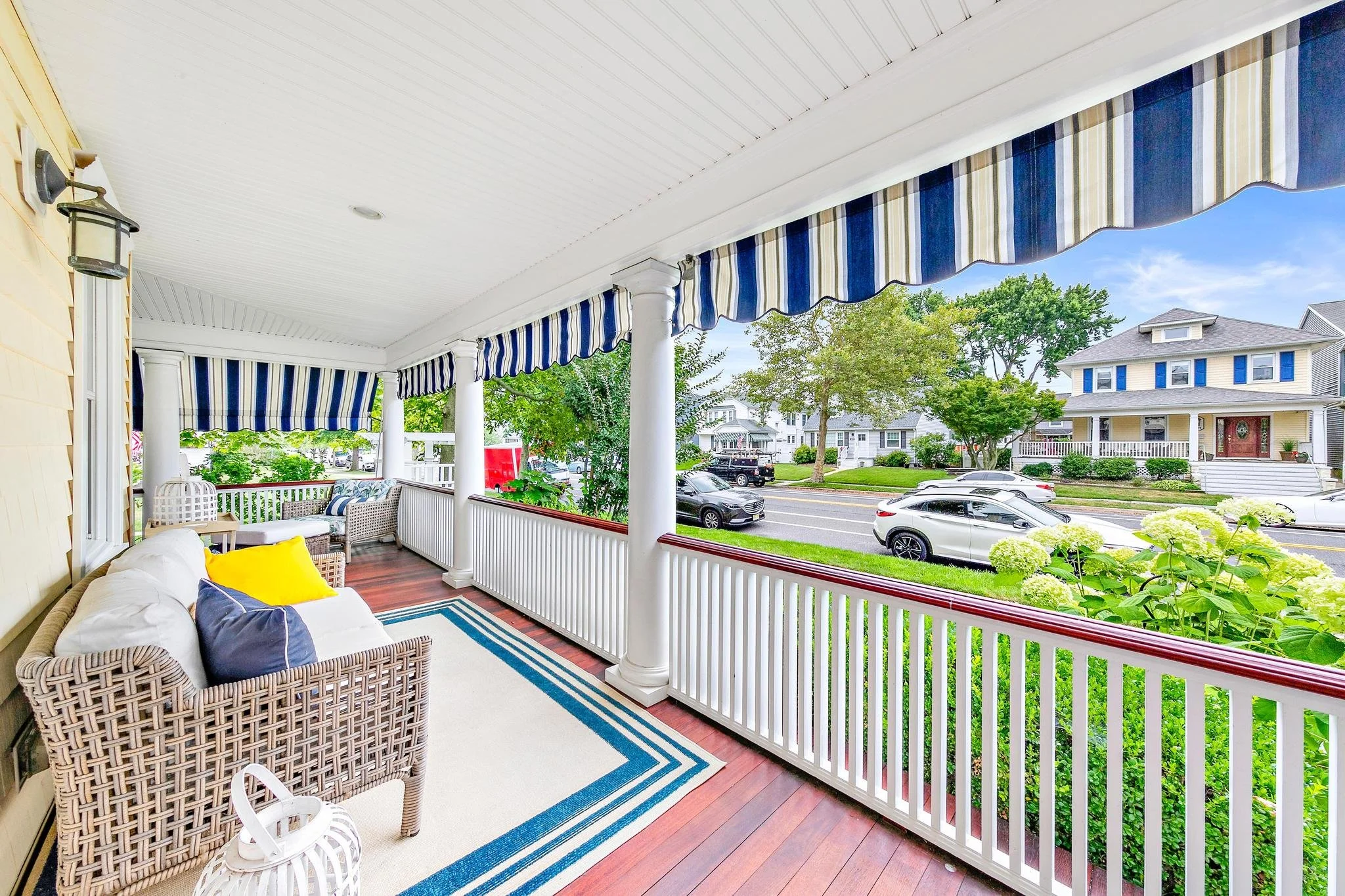 A spacious front porch with wicker and cushioned seating, striped blue and white awnings, white railings, and a view the trees and plantings around the home.
