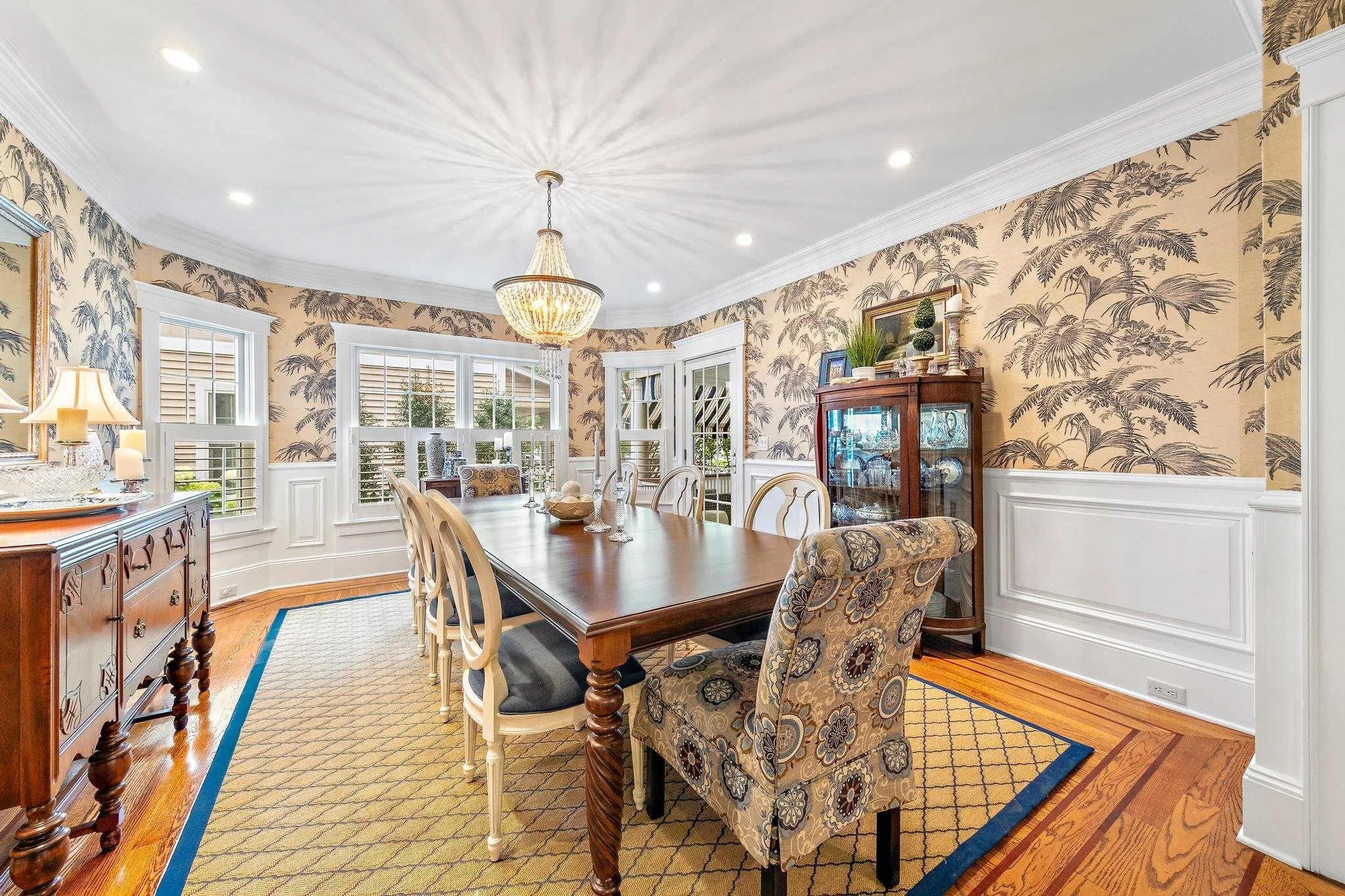 Elegant dining room with wood flooring, patterned wallpaper, a chandelier, a large wooden dining table, and various chairs. Windows with white trim and a china cabinet are also visible.