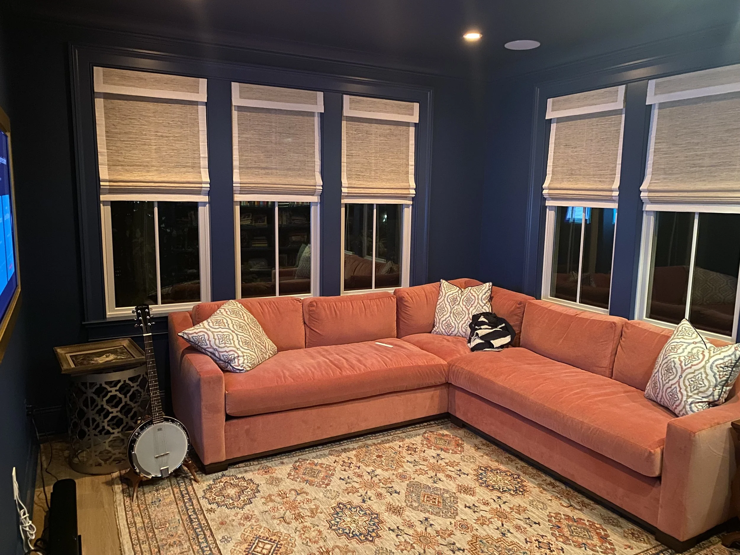 Den with a coral sectional sofa, three patterned pillows, a banjo, and an end table with a lattice design, against blue walls with four windows covered with beige roman shades, a sporting carpet, and a mounted television on the wall.