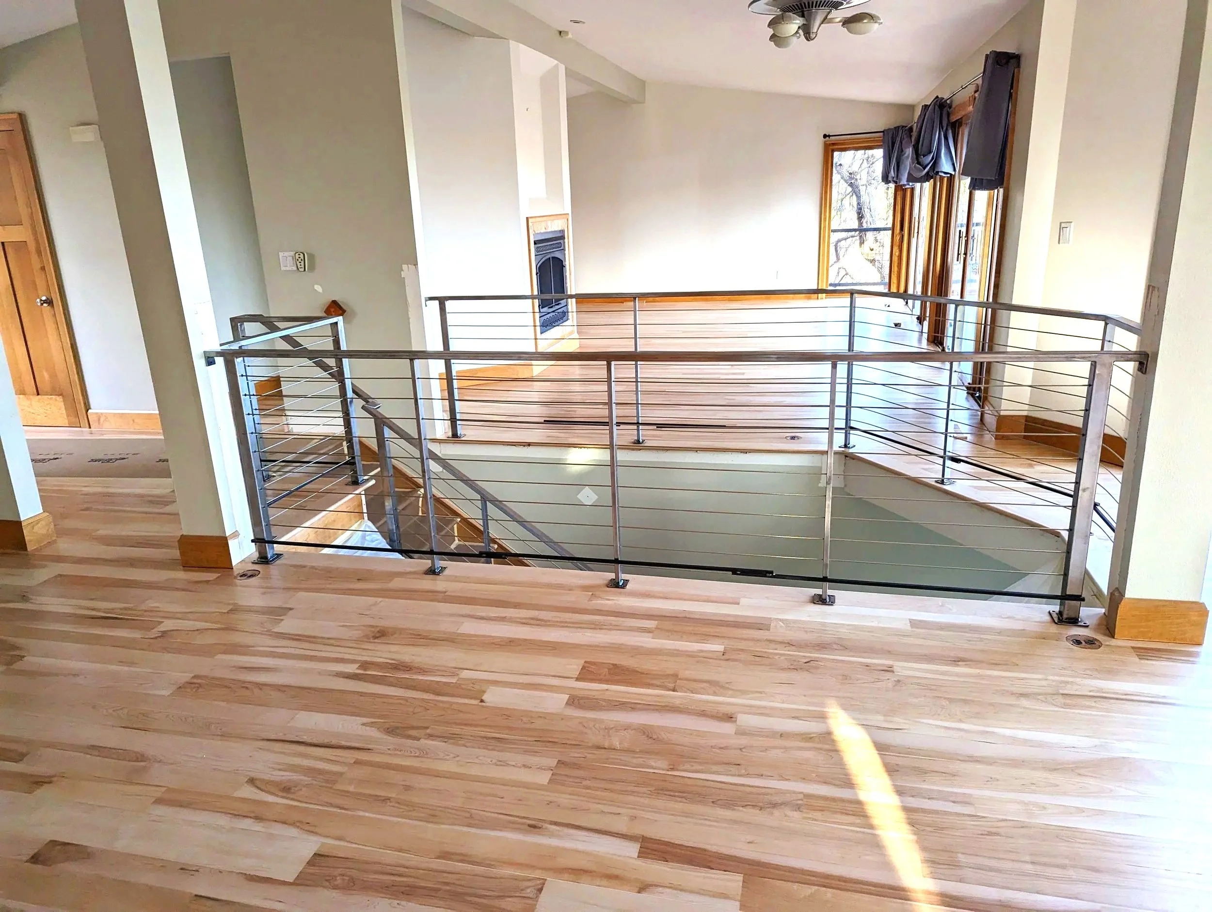 Interior view of a modern house with wooden floors, a staircase with metal railing, a fireplace, and large windows with curtains.