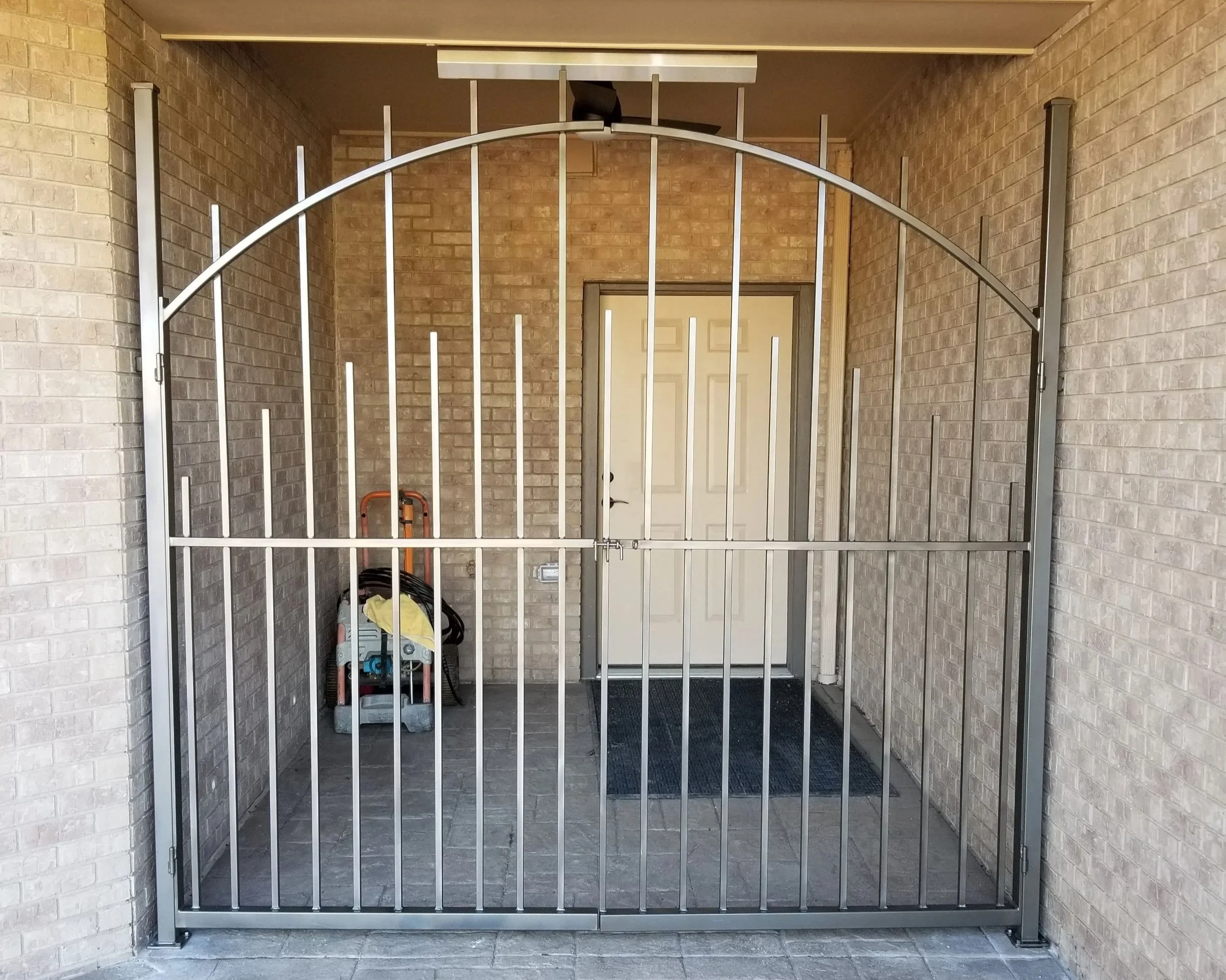 A metal gate blocking an entryway with a brick wall and a door behind it, a cleaning tool and a hose are visible on the left side of the entrance.