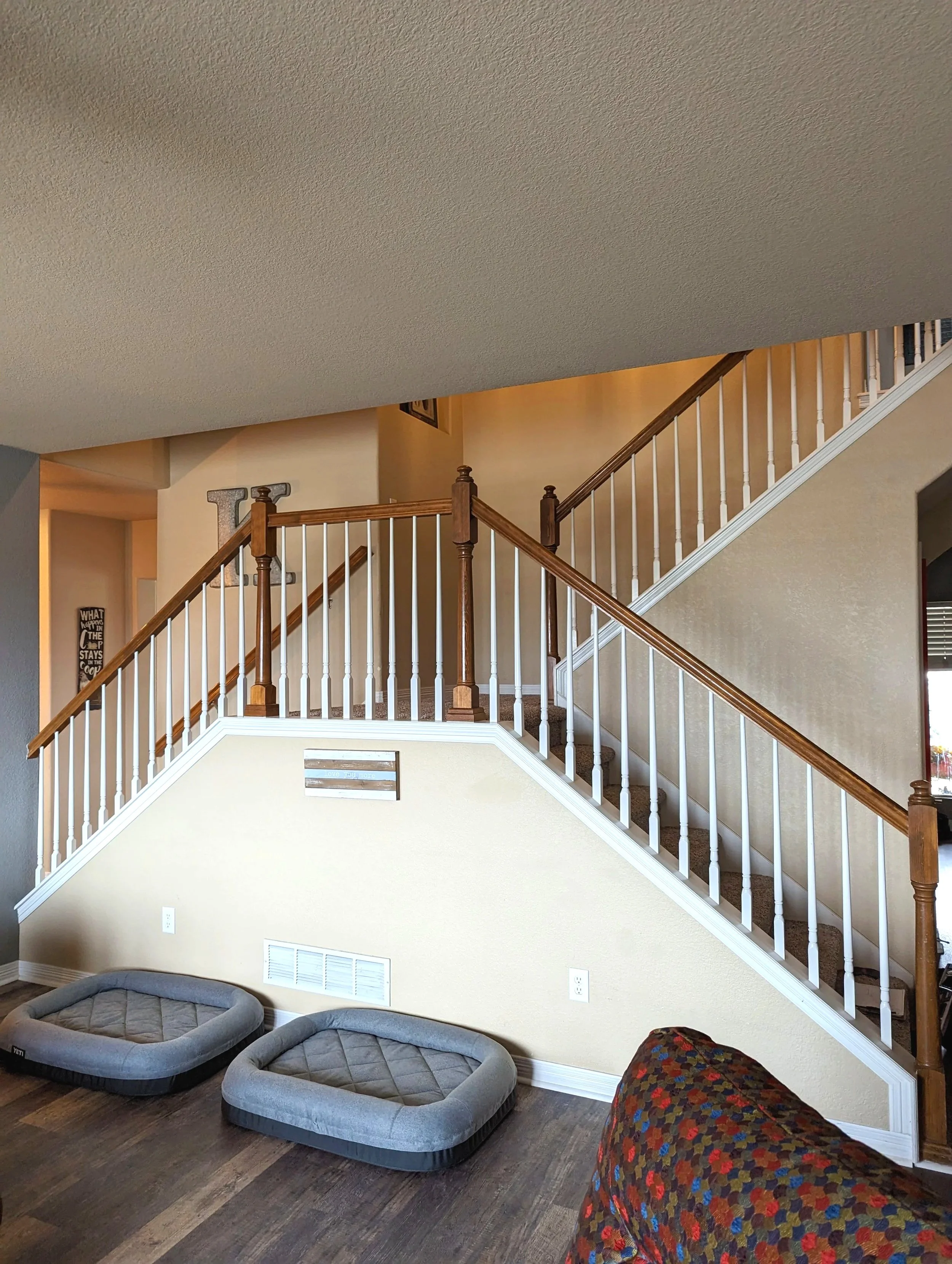 Living room with staircase, two grey pet beds on wood floor, beige wall, and a colorful chair with a floral pattern.