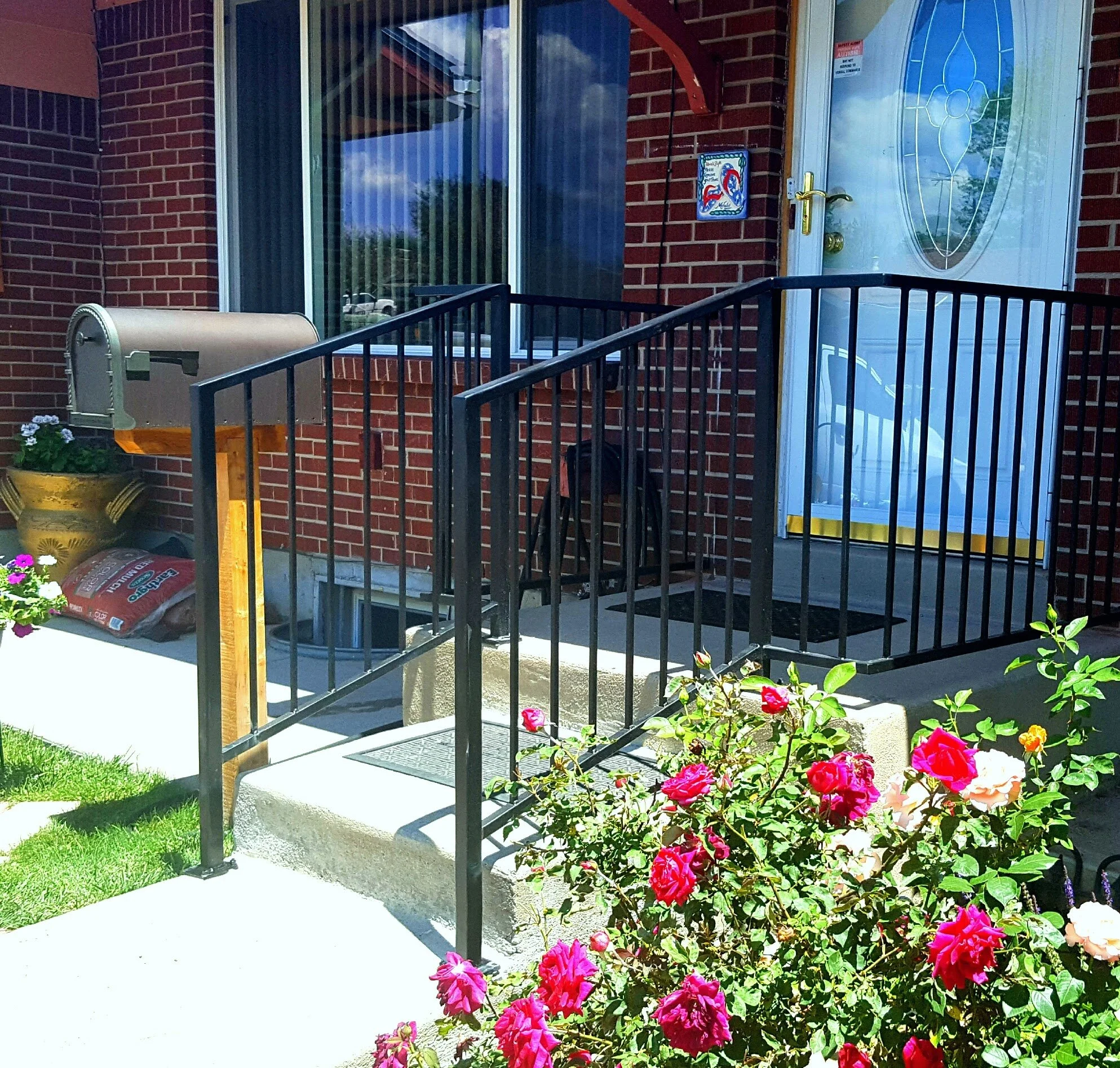 Front porch with black metal railing, brick house with glass front door, white storm door, and a window with vertical blinds. There is a mailbox, a potted plant with pink flowers, and a bag of mulch on the ground. Bright sunlight and greenery are visible.