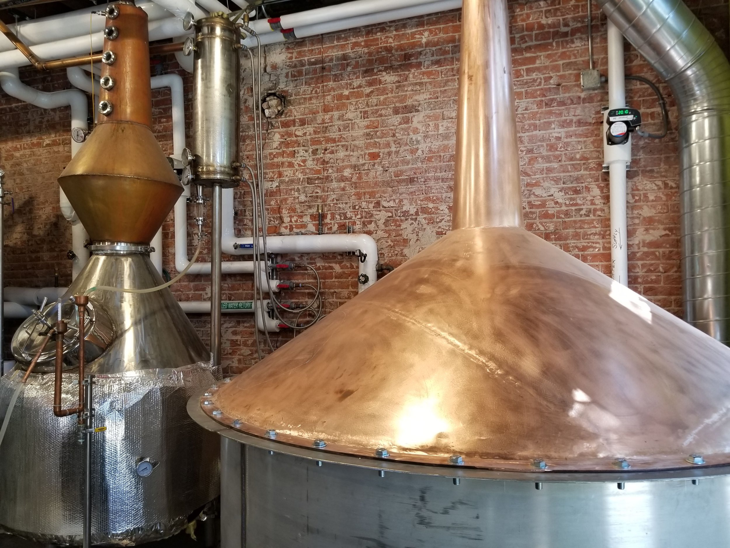 On-site Copper and  stainless steel metal distillation equipment in a distillery with pipes, gauges, and a brick wall background.