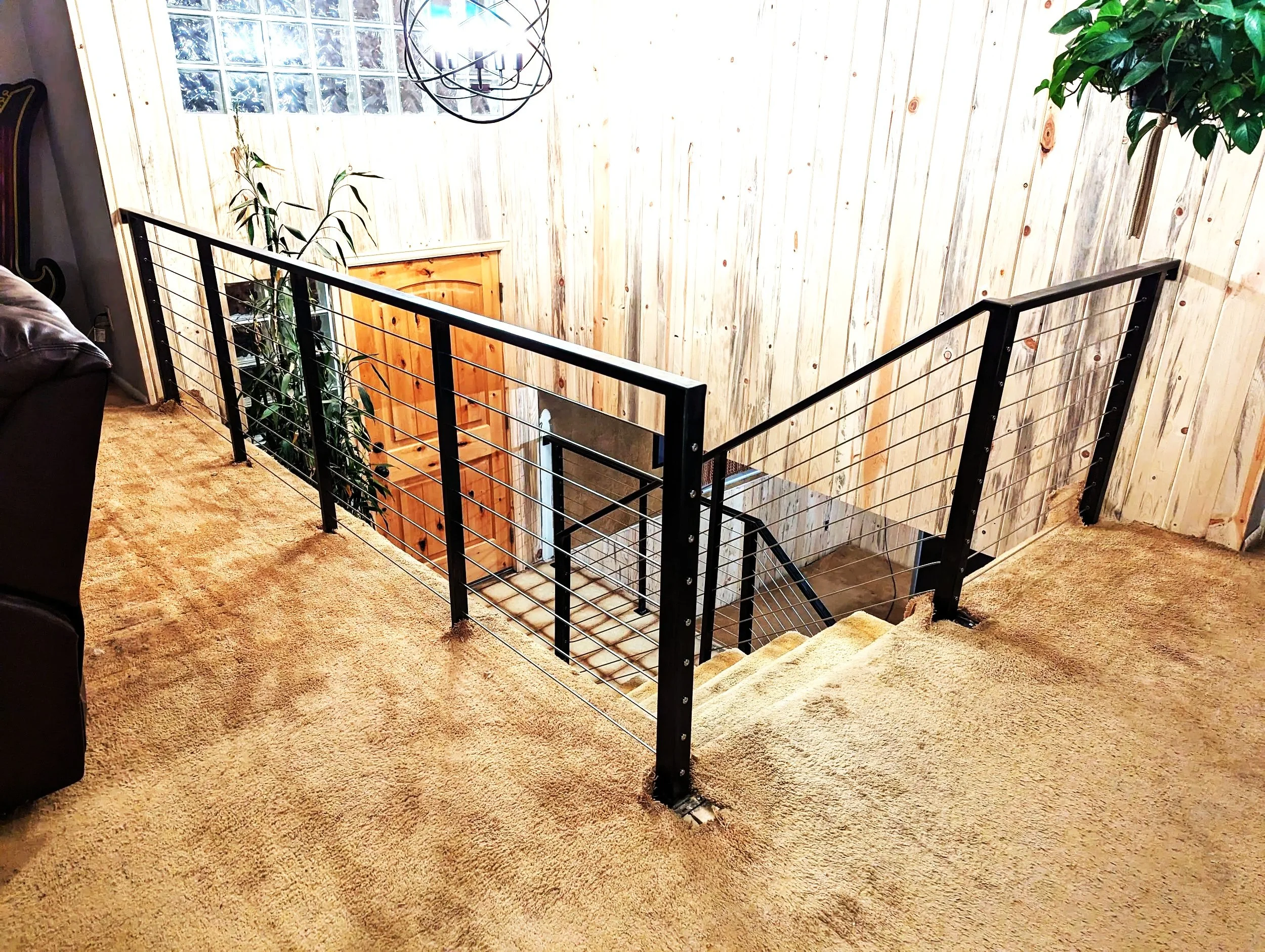 Interior view of a home staircase landing with a wooden door, a black metal railing, plants, a chandelier, and carpeted stairs.
