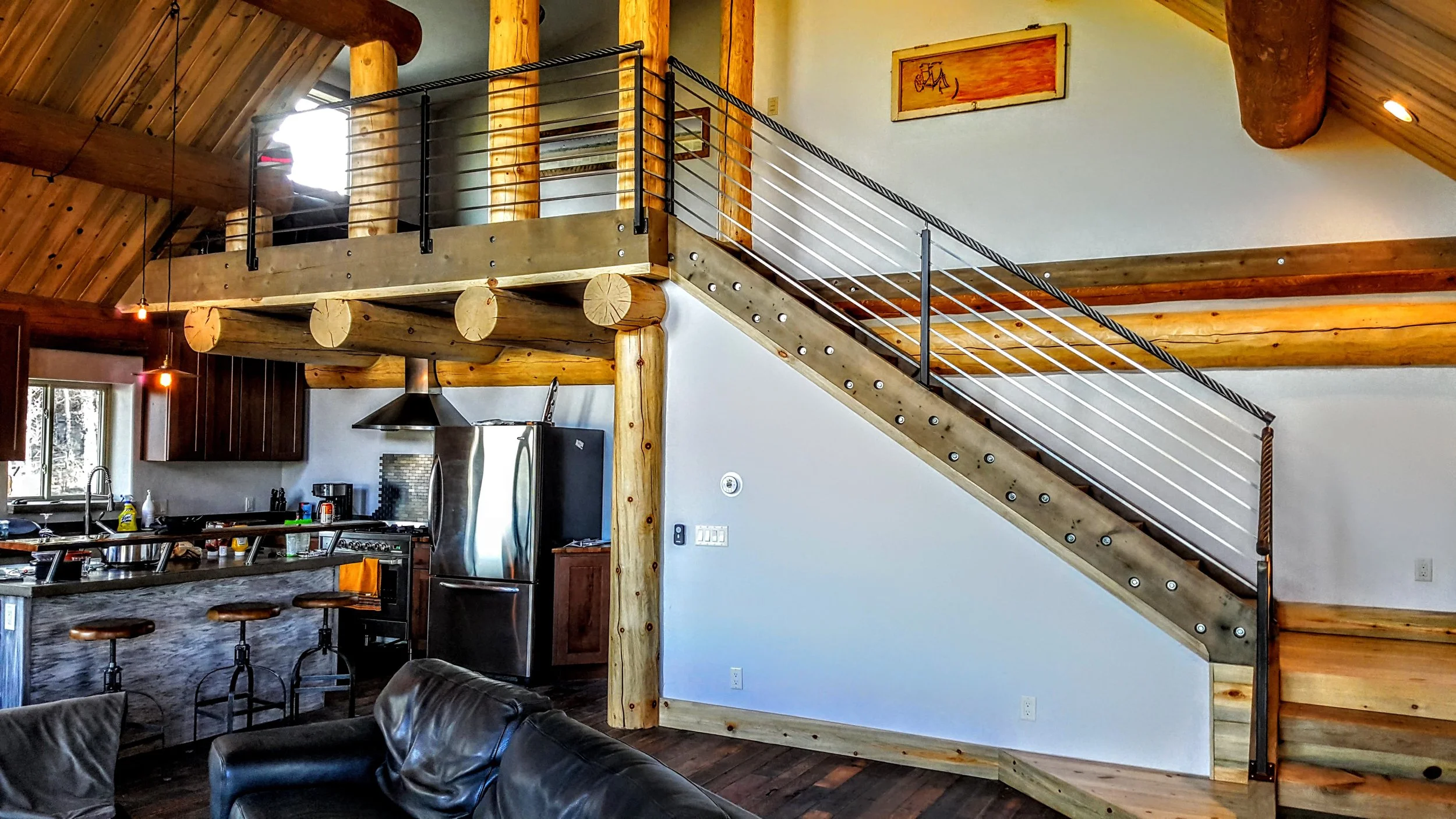 Metal railing with a custom ski cable handrail in a rustic cabin house with kitchen area with dark wooden cabinets and stainless steel fridge.