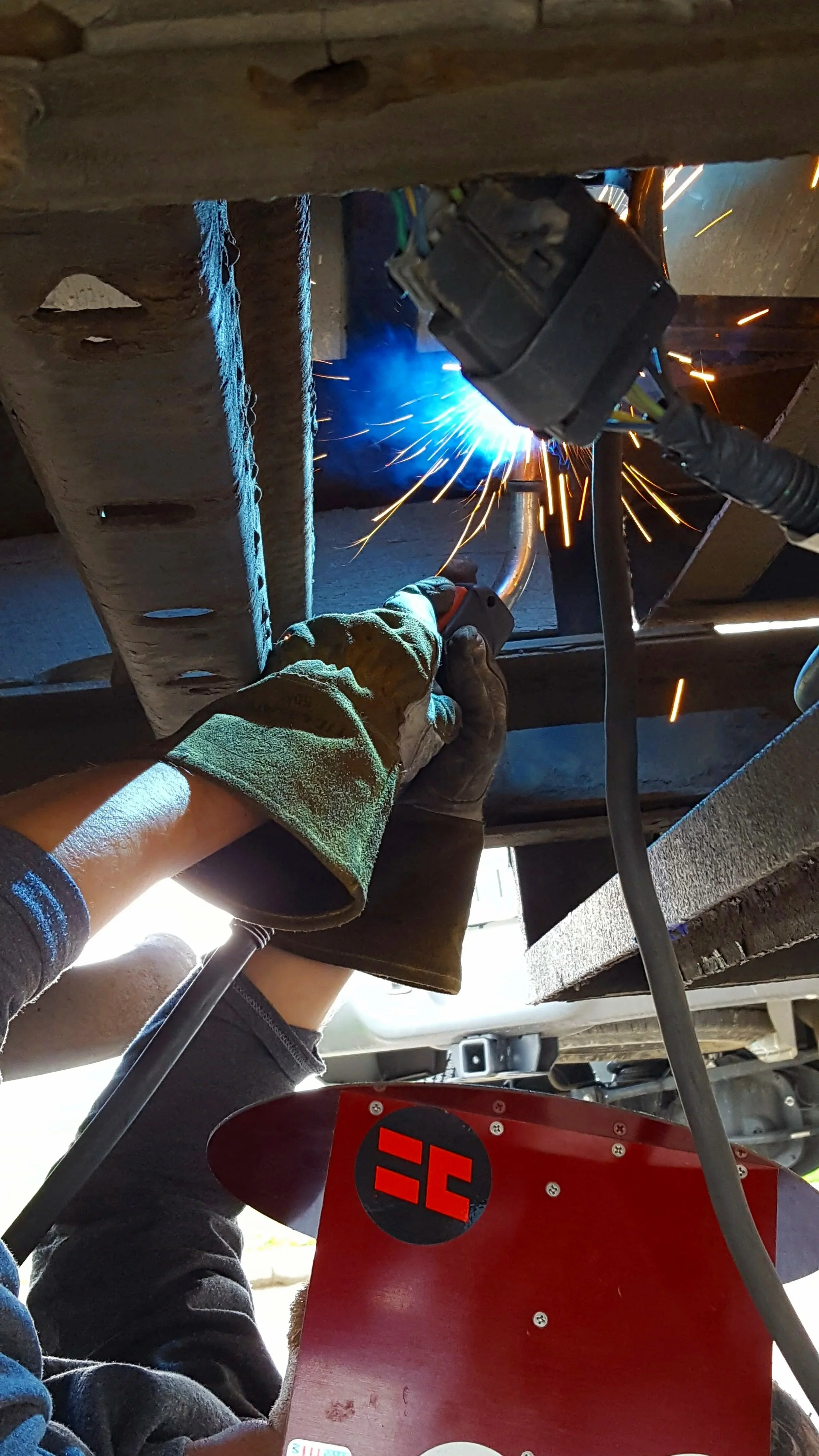 A person welding underneath a vehicle, sparks flying, wearing gloves and a protective sleeve.