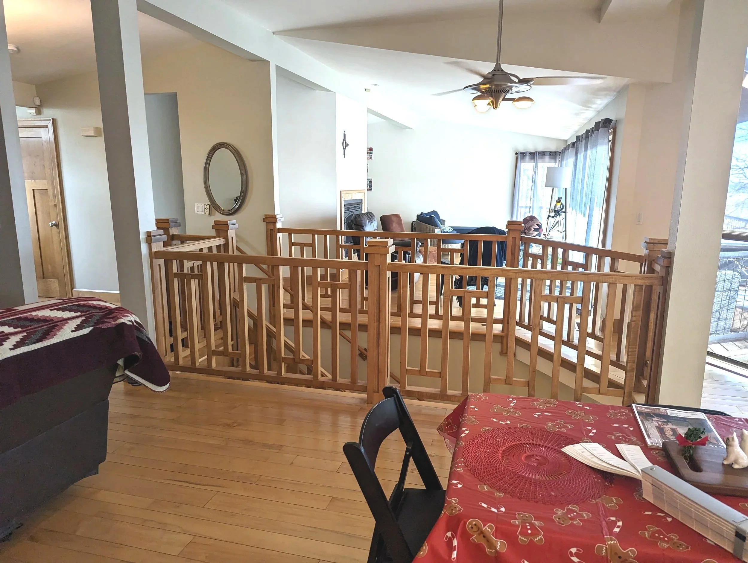 Living room with a staircase, a dining table with a red holiday-themed tablecloth, small holiday decorations, and a sitting area with chairs and a lamp near large windows with curtains.