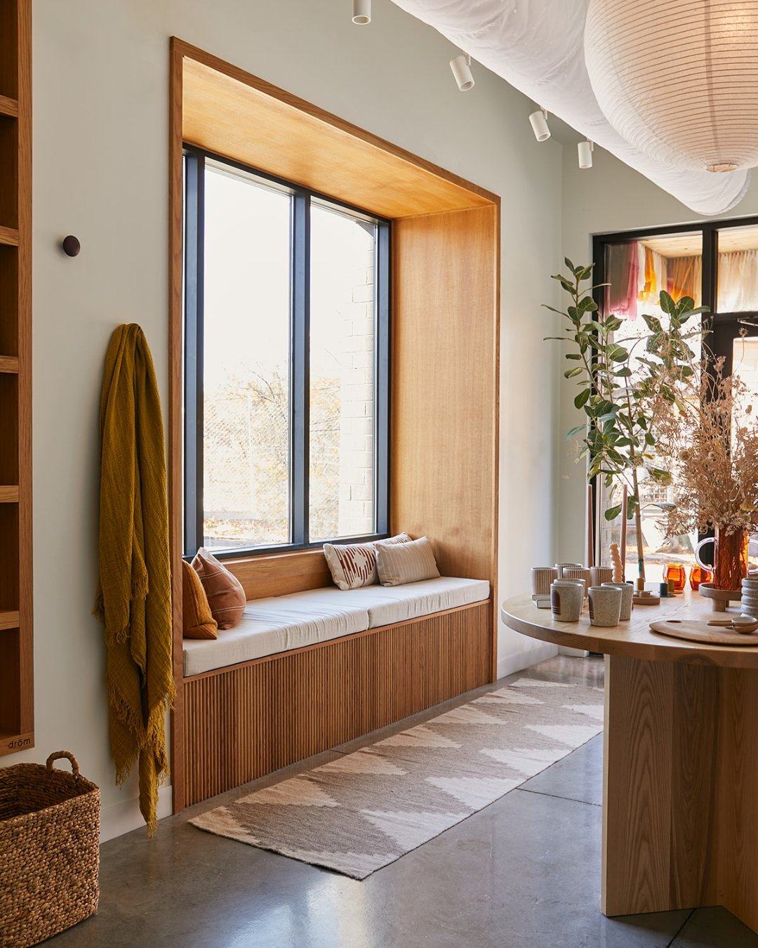 Cozy sunlit window nook with a cushioned bench, pillows, and a view of trees outside. Modern interior decor with a wooden accent wall and table, potted plants, and a variety of cups and decorative items.