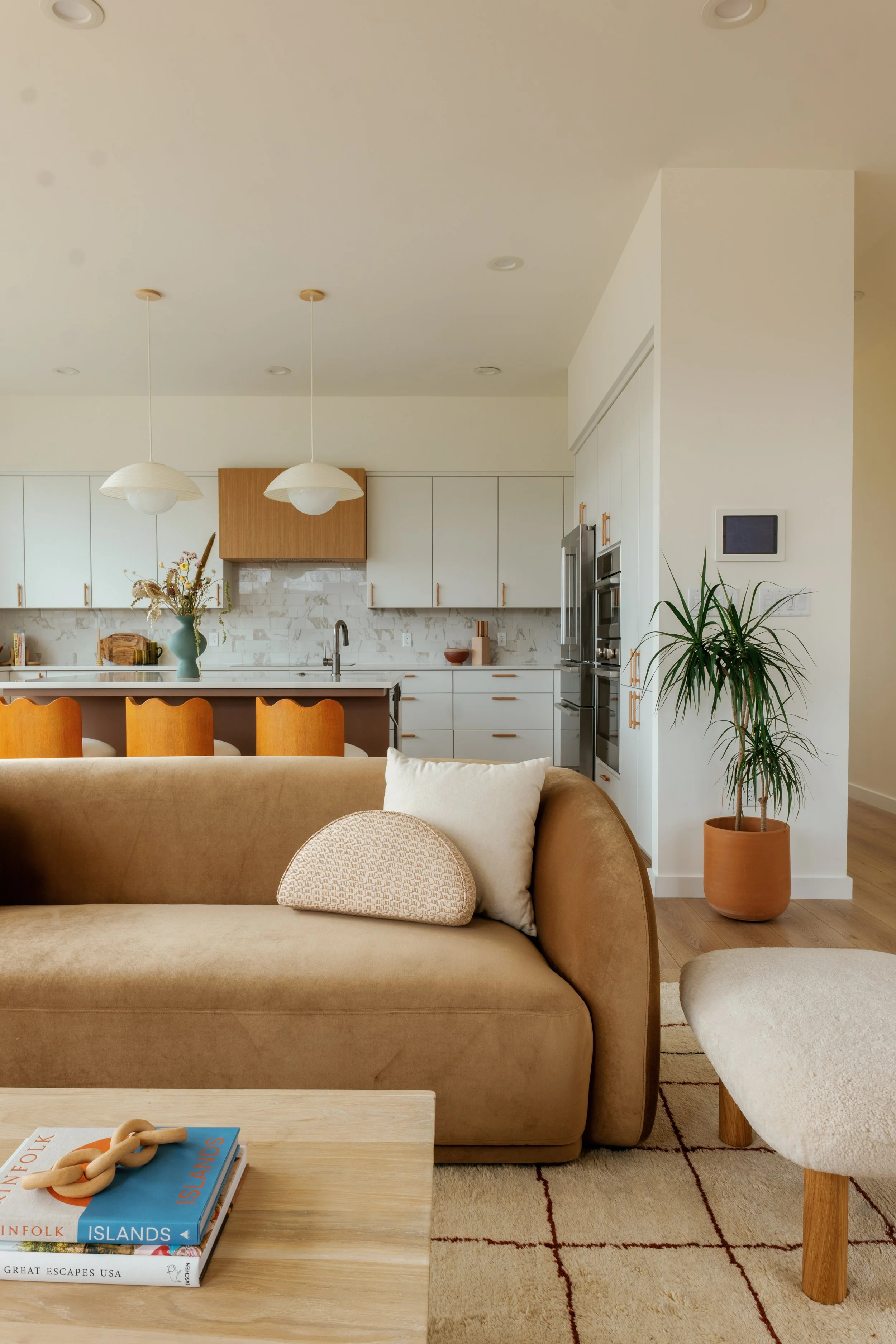 Open-concept living room with beige sofa, white pillows, wooden coffee table with books, and a nook with a kitchen in the background, including white cabinetry, a marble backsplash, hanging pendant lights, a vase with flowers, and a potted plant.