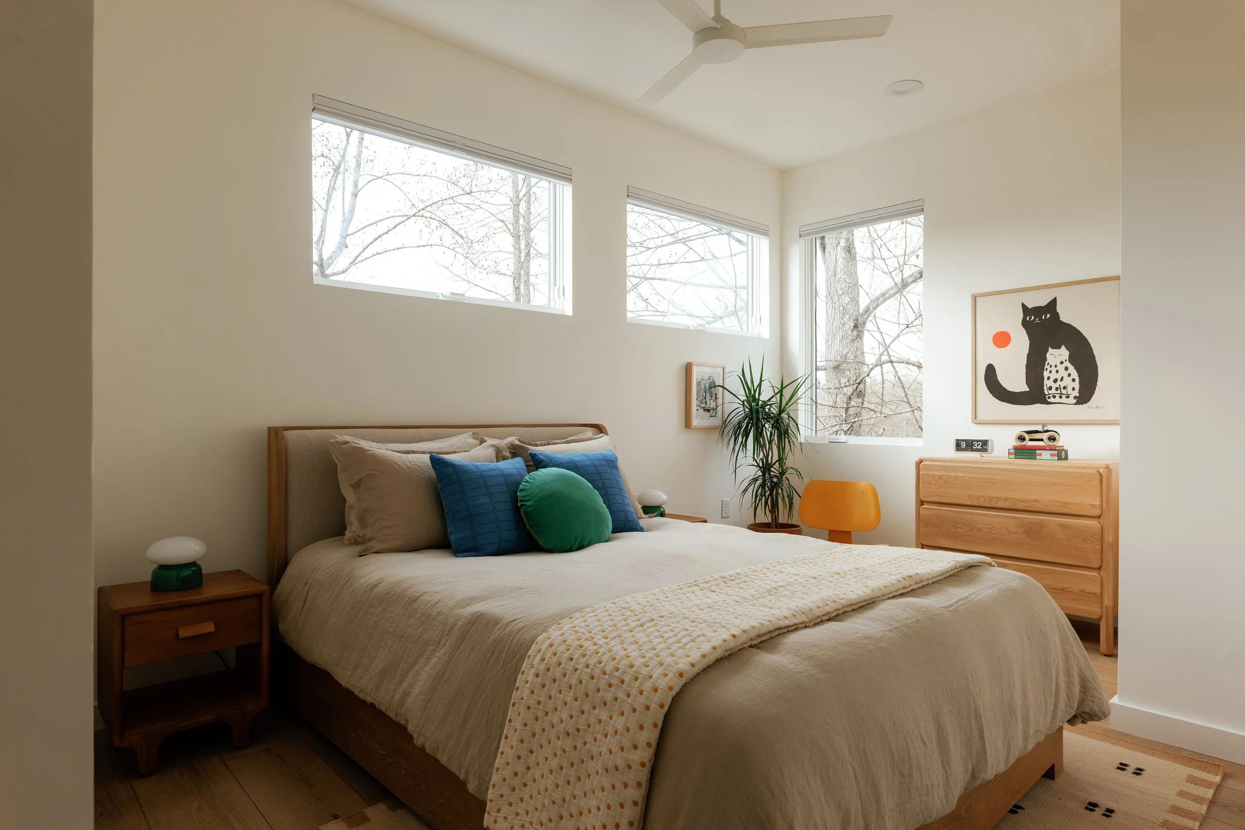 A bedroom with white walls and a wooden bed frame. The bed has beige sheets, gray and blue pillows, and a green round pillow. There are two wooden nightstands with small lamps. A large window shows leafless trees outside. A yellow chair, a potted plant, and a wooden dresser with artwork of a black cat and a white spotted cat are present.