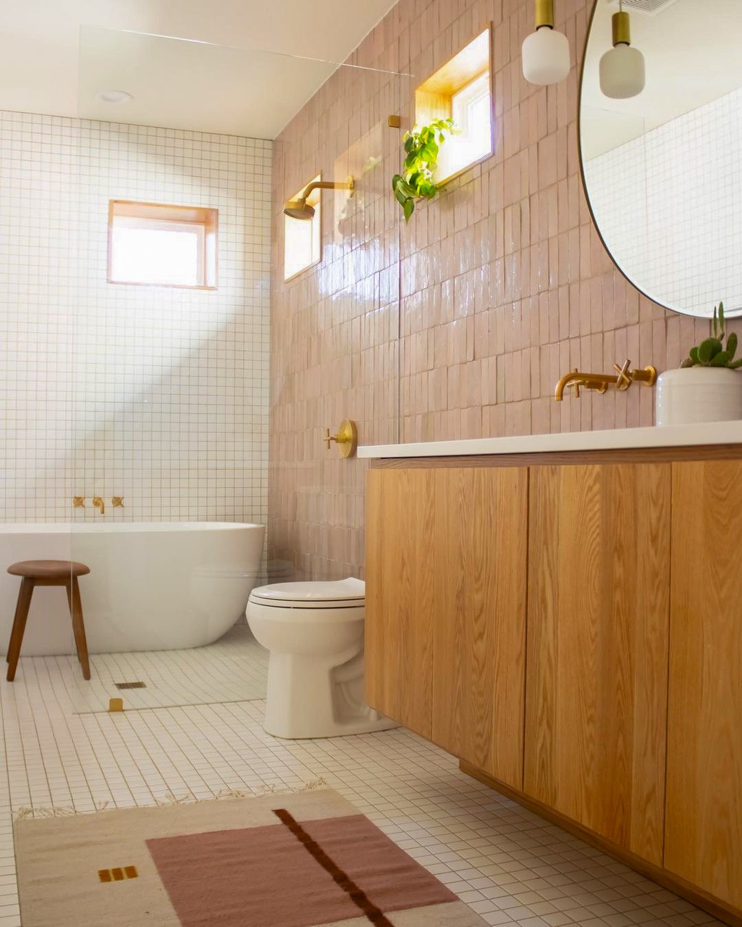 Bathroom with shower, toilet, wooden vanity with gold fixtures, small stool, round mirror, window with a plant, and tiled walls and floor.
