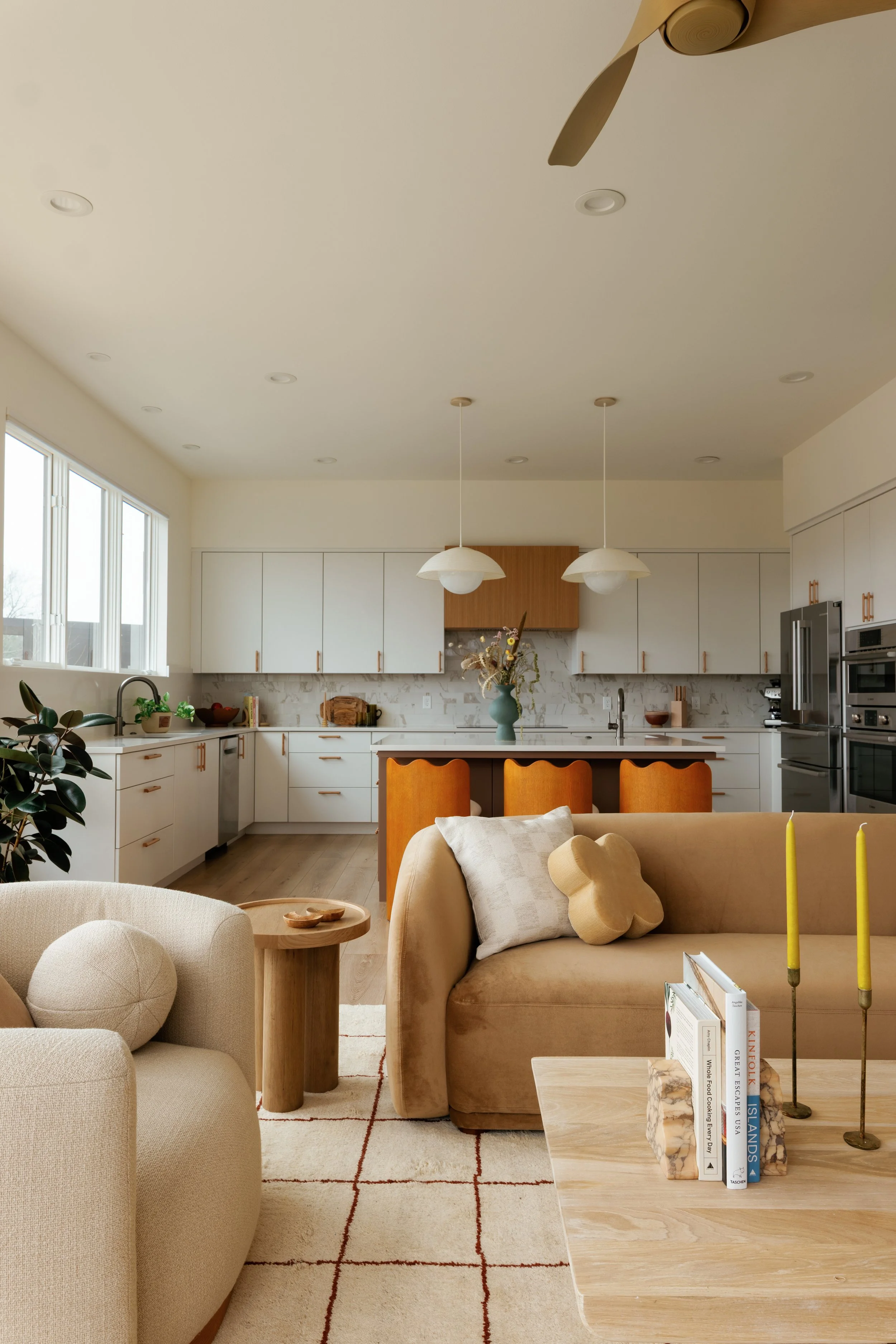 Bright, open-concept kitchen and living room with white cabinets, wooden accents, beige sofas, a coffee table with books, and large windows.