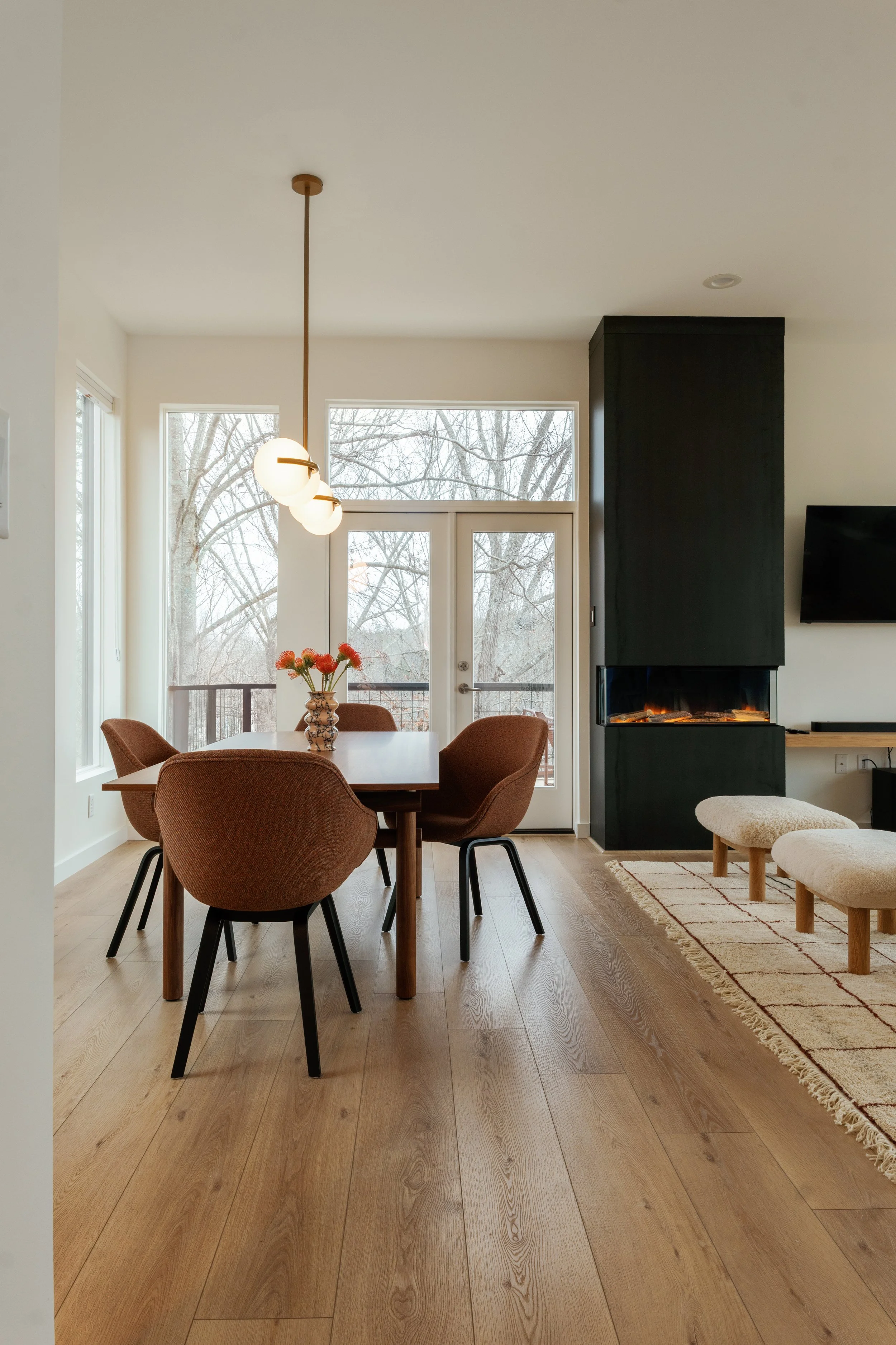 A room with a round wooden dining table surrounded by four upholstered brown chairs, a vase with orange flowers, a black fireplace with a fire inside, a large window and glass door showing trees outside, and a hanging light fixture with three globe bulbs.
