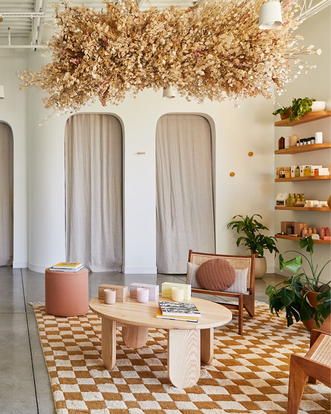 A cozy, modern interior with beige curtains, a round wooden coffee table, a pink ottoman, rattan chairs with cushions, and potted plants. There are wooden shelves on the right filled with various products, and a large floral arrangement hanging from the ceiling.