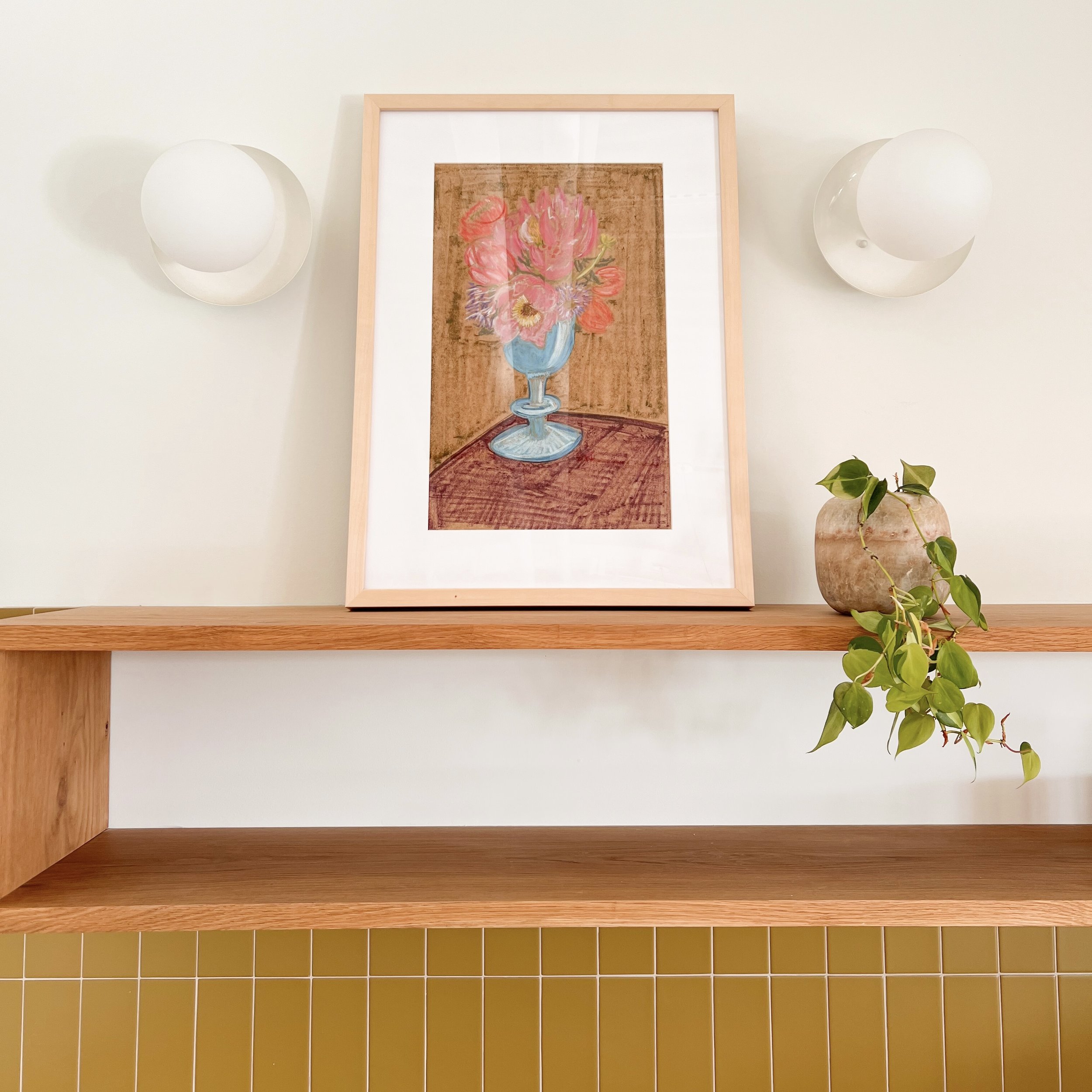 A framed painting of pink and red flowers in a blue vase on a wooden table, mounted on a white wall with two modern wall sconces and a potted plant on a wooden shelf underneath.