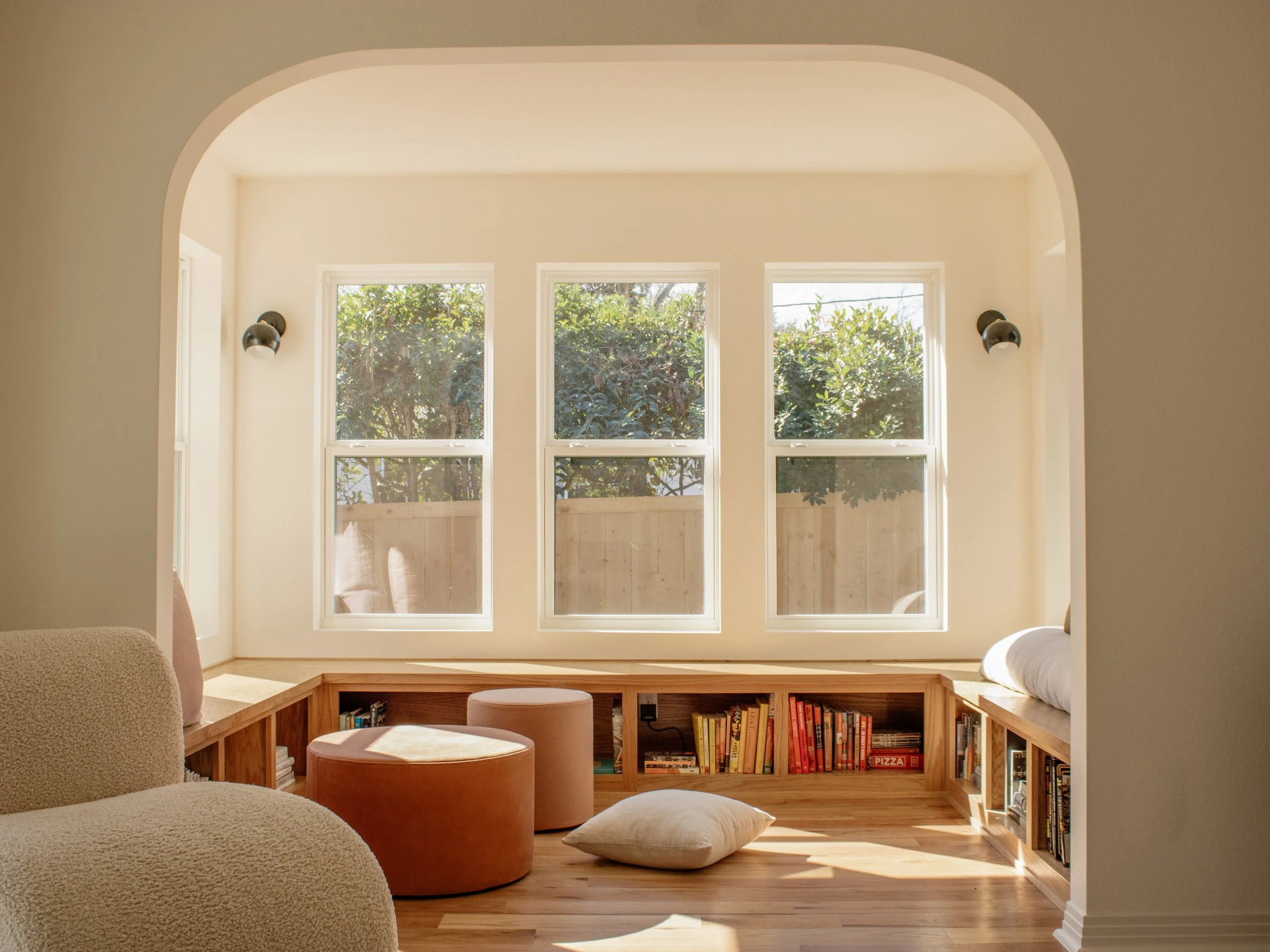 Sunlit cozy reading nook with built-in wooden bookshelf beneath large windows, cushioned seating, a pillow on the floor, and a view of trees outside.
