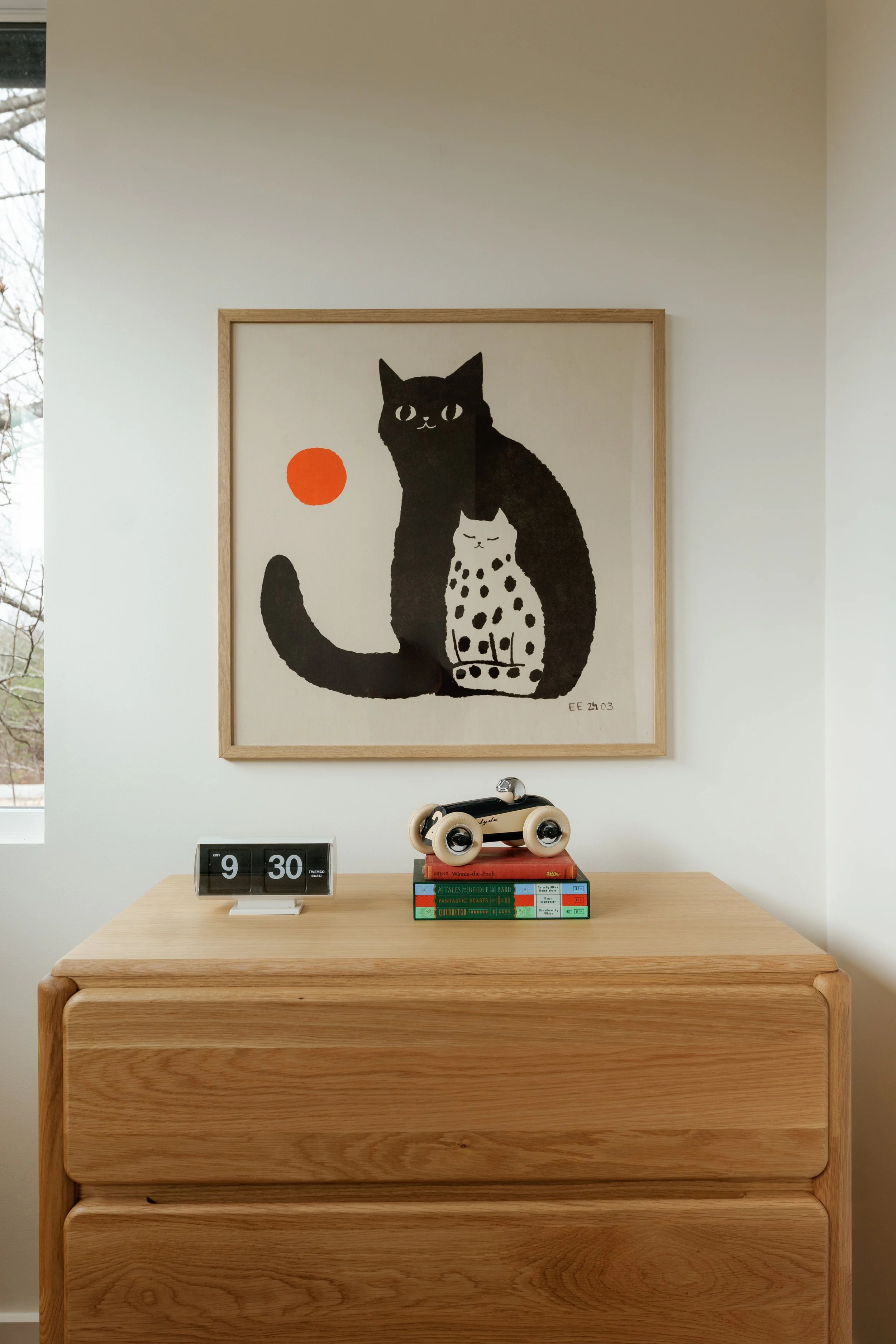 Decorative wooden dresser with a vintage tandem roller skate and books on top, framed artwork of two stylized black and white cats with a red circle on a white wall behind.