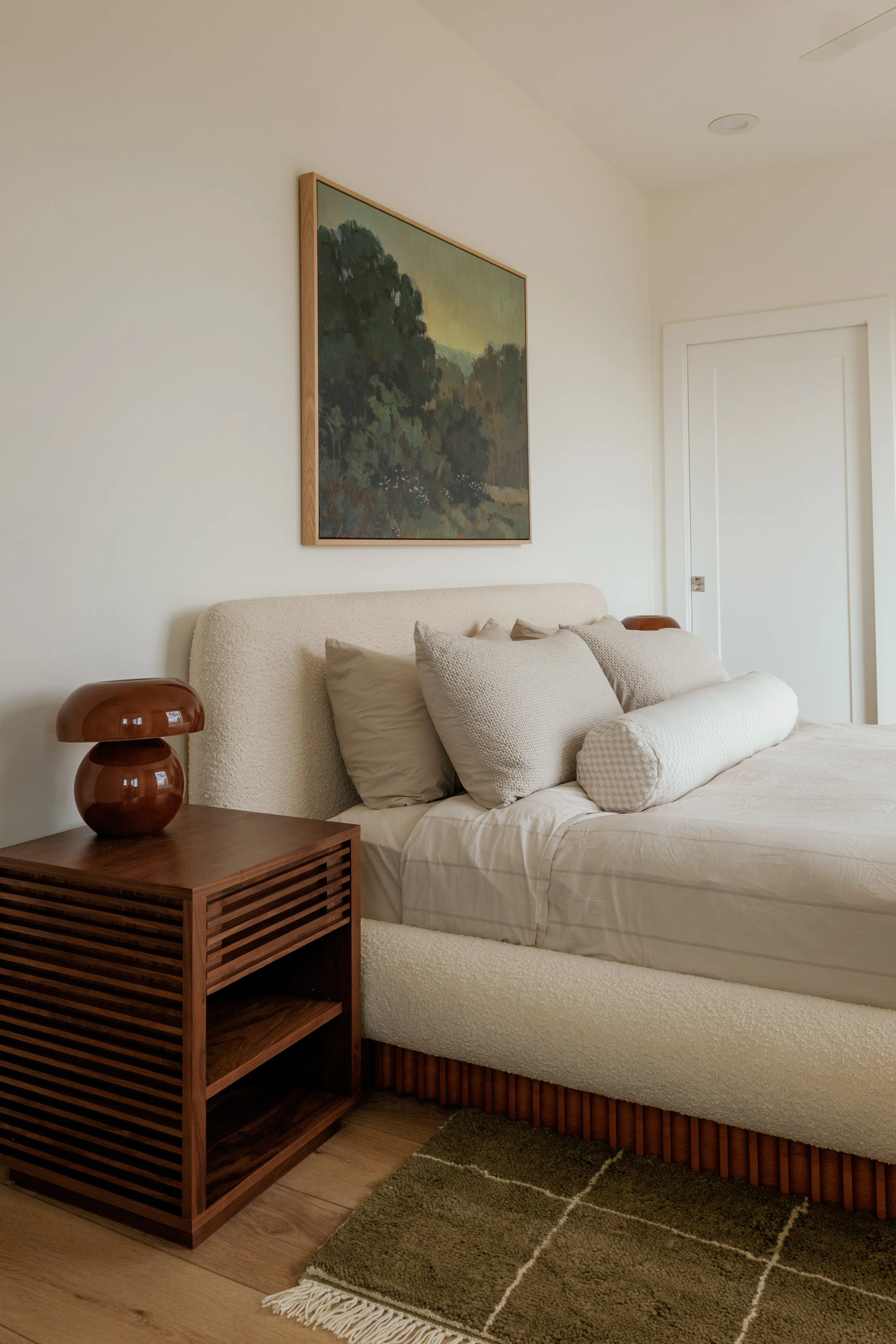 A bedroom with a beige upholstered bed, a wooden nightstand with a brown ceramic table lamp, a landscape painting above the bed, closed white door, beige and gray pillows, and a green rug on a wooden floor.