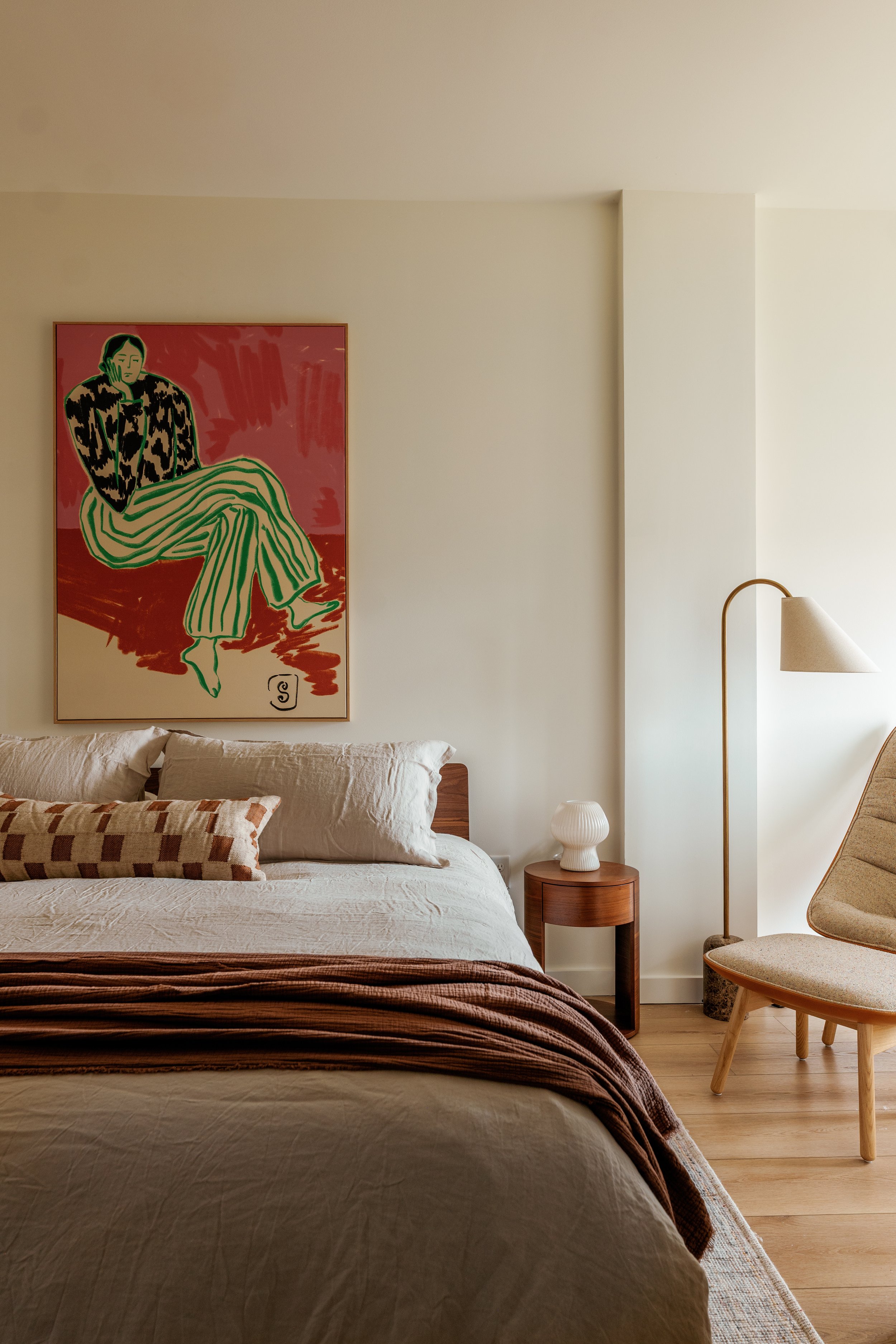 A bedroom with a bed, beige pillow, and a blanket, a small round wooden side table with a white vase, an armchair with light upholstery, a modern floor lamp, and a colorful artwork of a woman sitting with striped green and beige pants on the wall.