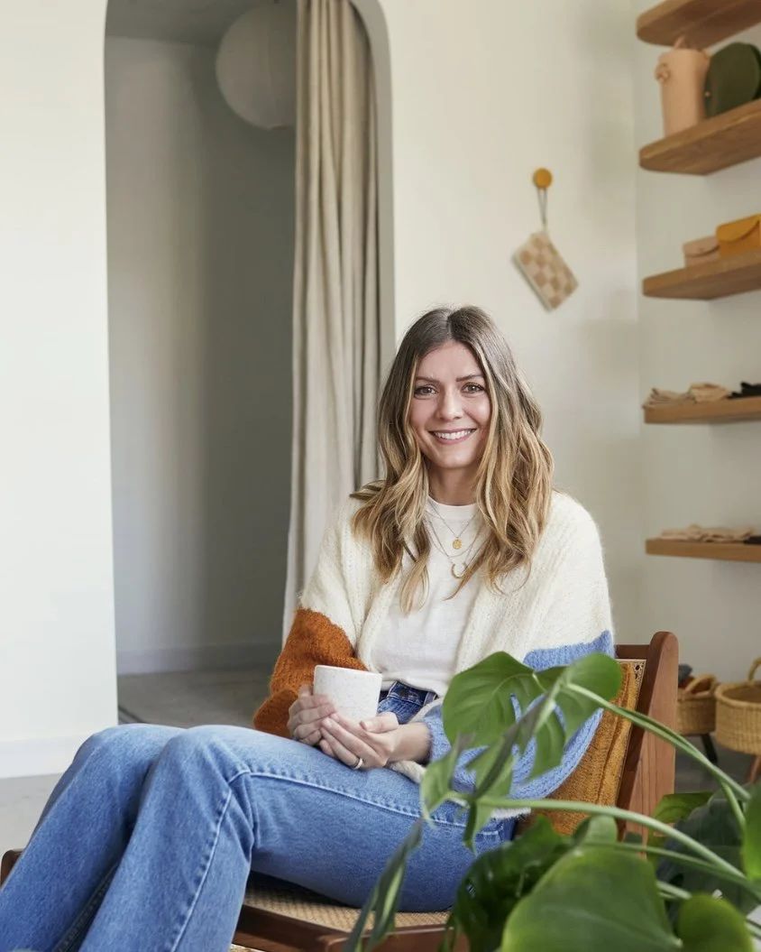 A woman with wavy, shoulder-length hair smiling and sitting in a cozy room, holding a white mug, with green houseplants and wooden shelves in the background.