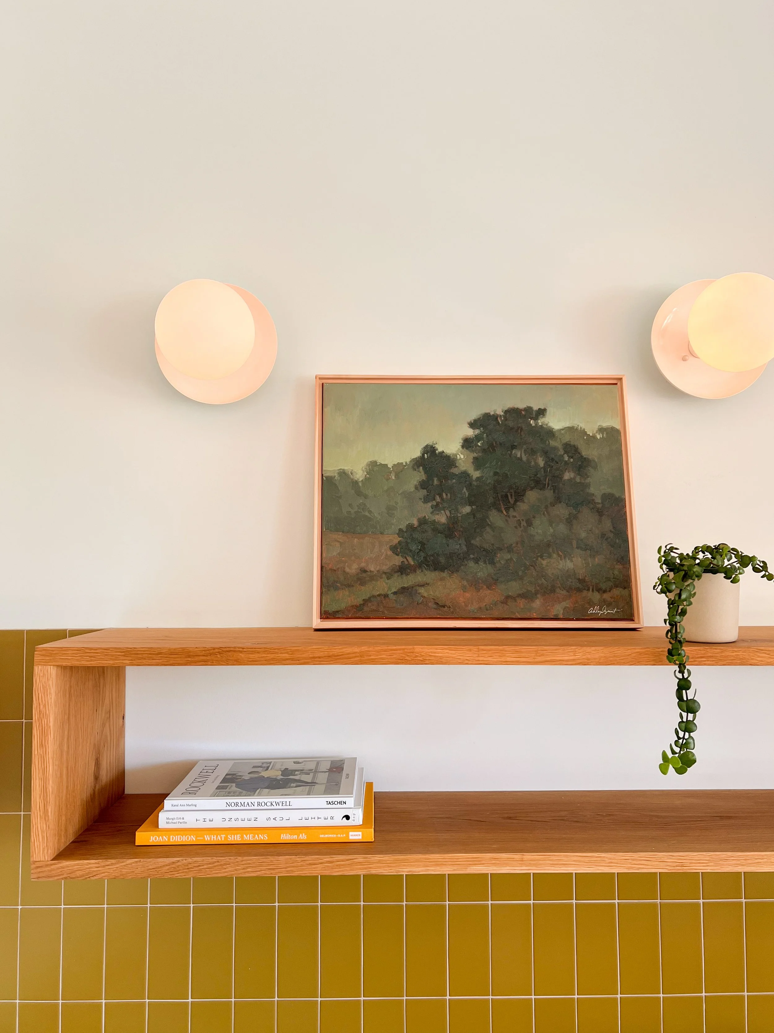 A wooden shelf with a framed landscape painting of trees, a small potted plant with trailing greenery, and several books, set against a white wall with two round wall sconces.