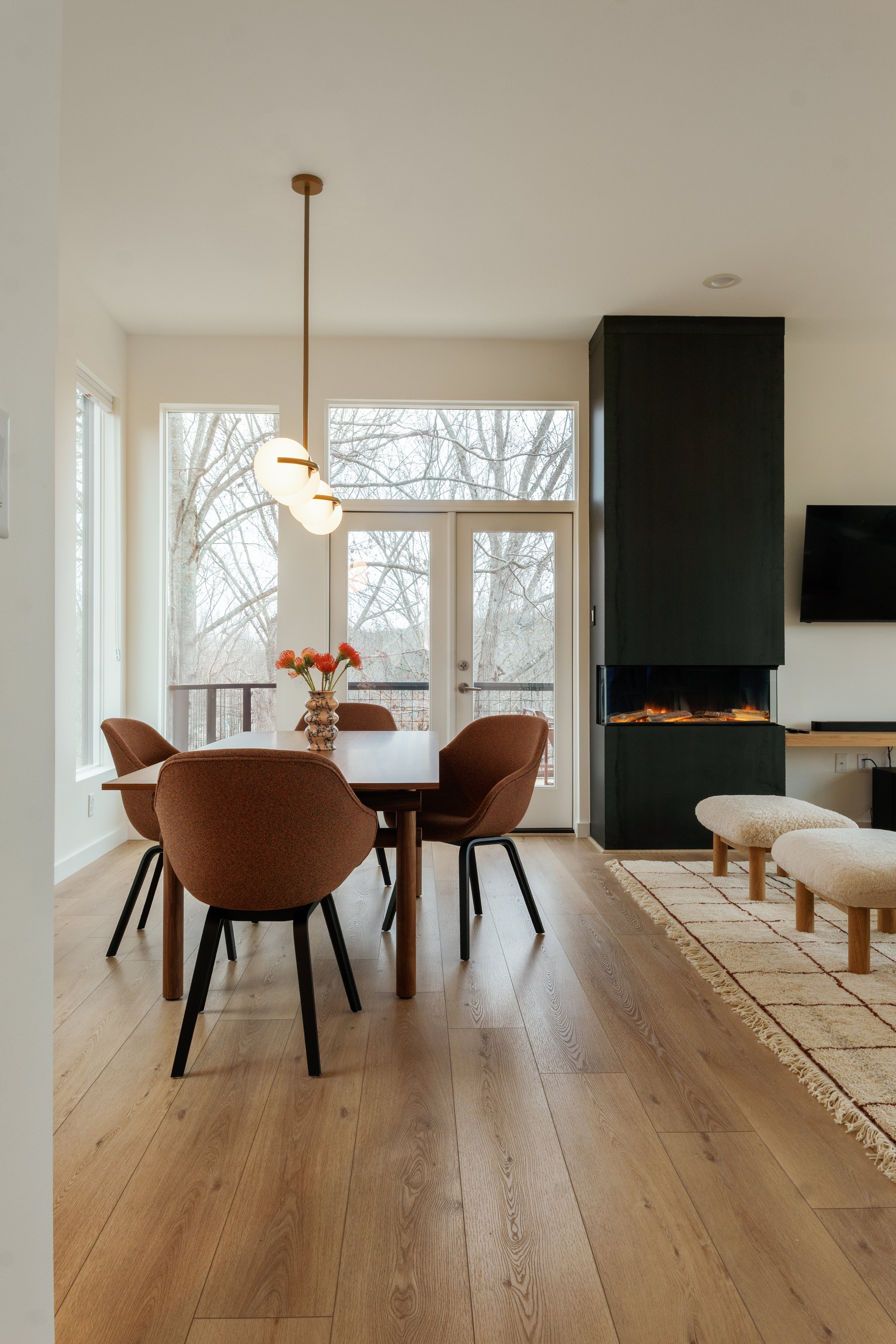 Interior of a modern dining and living area with a wooden dining table, six brown upholstered chairs, a hanging light fixture, a fireplace, a flat-screen TV, and a beige area rug on wooden flooring, with large windows and a door leading outside.
