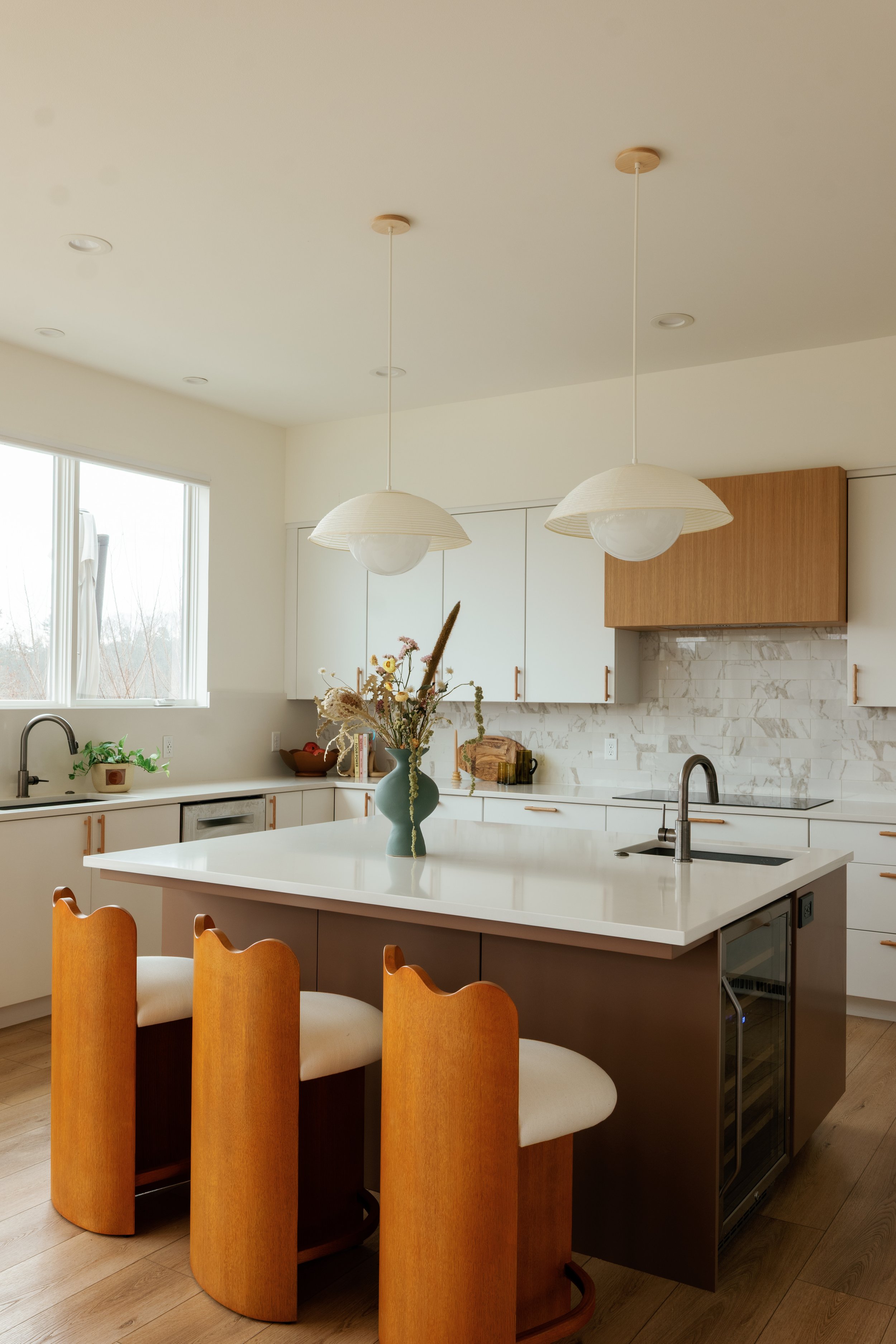 Modern kitchen with a white island, wooden chairs, white cabinets, a marble backsplash, and pendant lights.