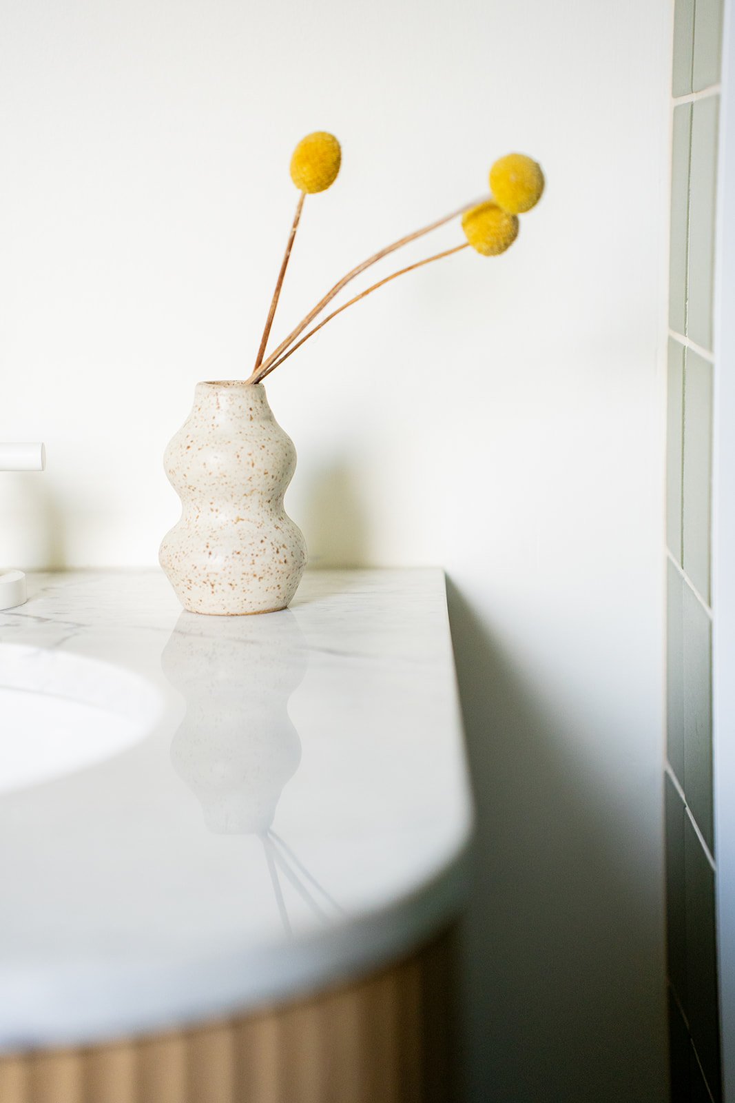 A beige vase with a textured surface and curvy shape holding three yellow billy ball flowers on a white marble countertop, with a plain light-colored wall in the background.