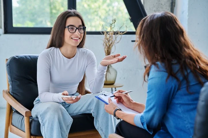 A young woman wearing glasses and a white long-sleeve shirt sitting on a black cushioned chair, smiling and holding a smartphone, talking to another woman with reddish-brown hair wearing a blue jacket, who is taking notes on a clipboard in an office with a large window and a plant in the background.