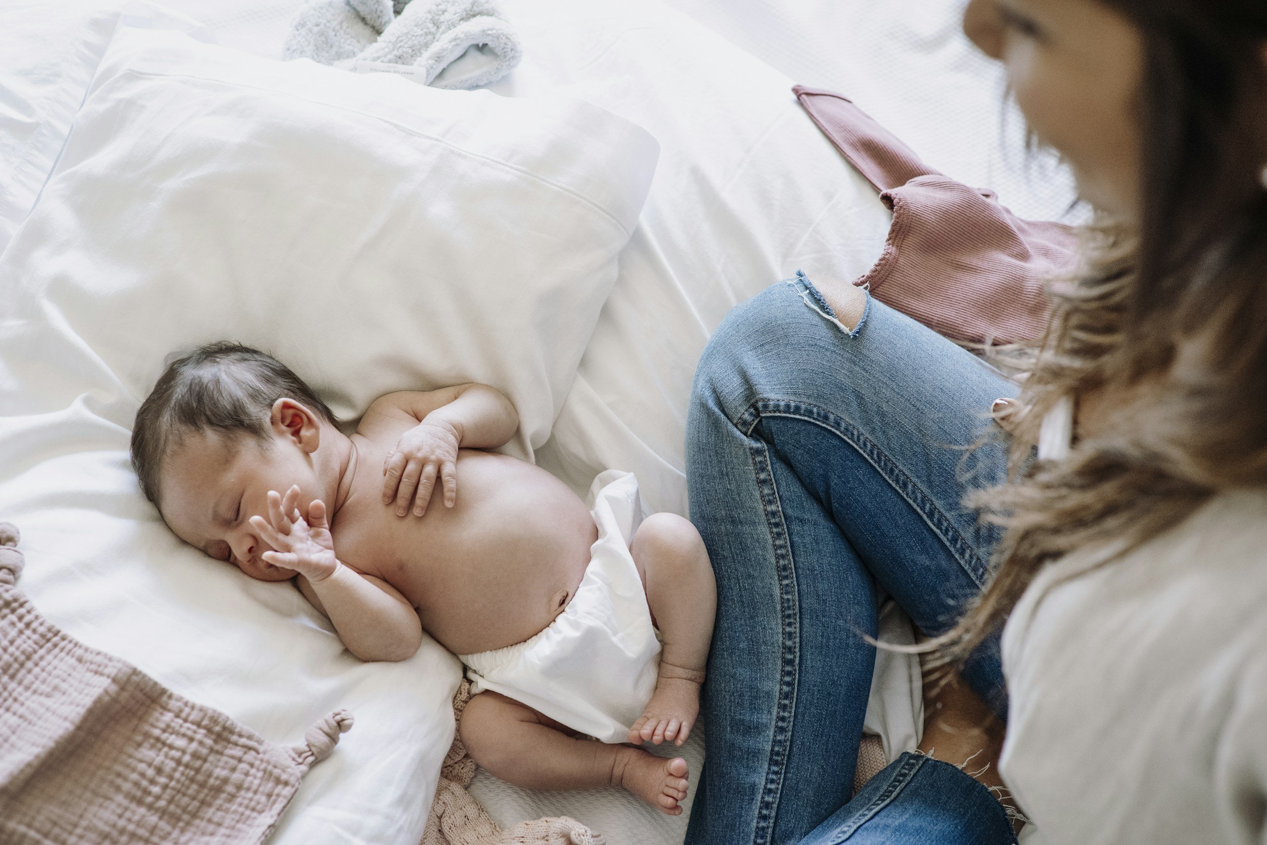 A newborn baby sleeping on a bed next to a woman, possibly his mother, who is sitting with her knees up, wearing jeans and a pink top.