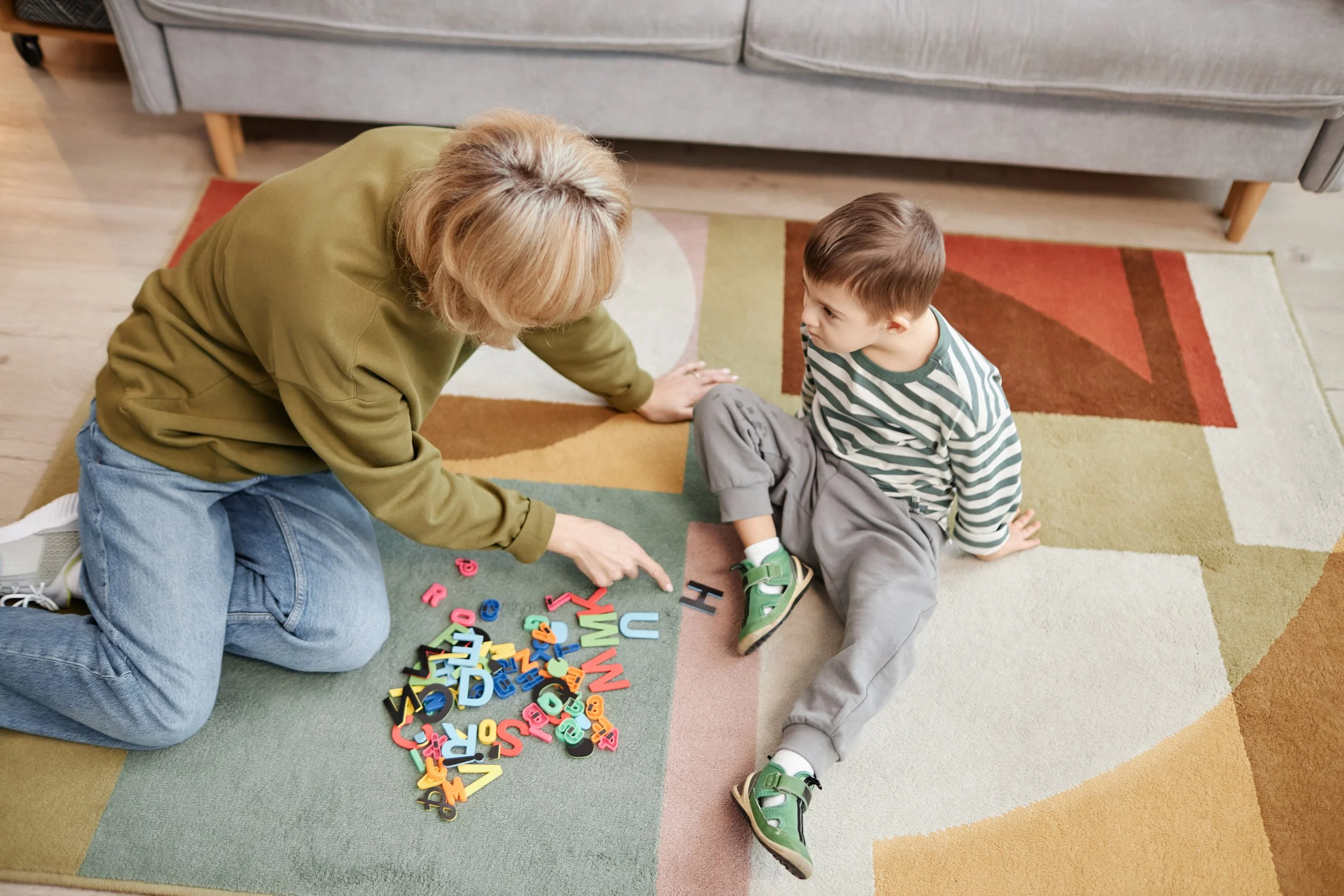 A woman and a young boy sitting on a multicolored rug, playing with colorful alphabet magnets. The woman is reaching for the magnets while the boy looks at her, sitting with crossed legs.
