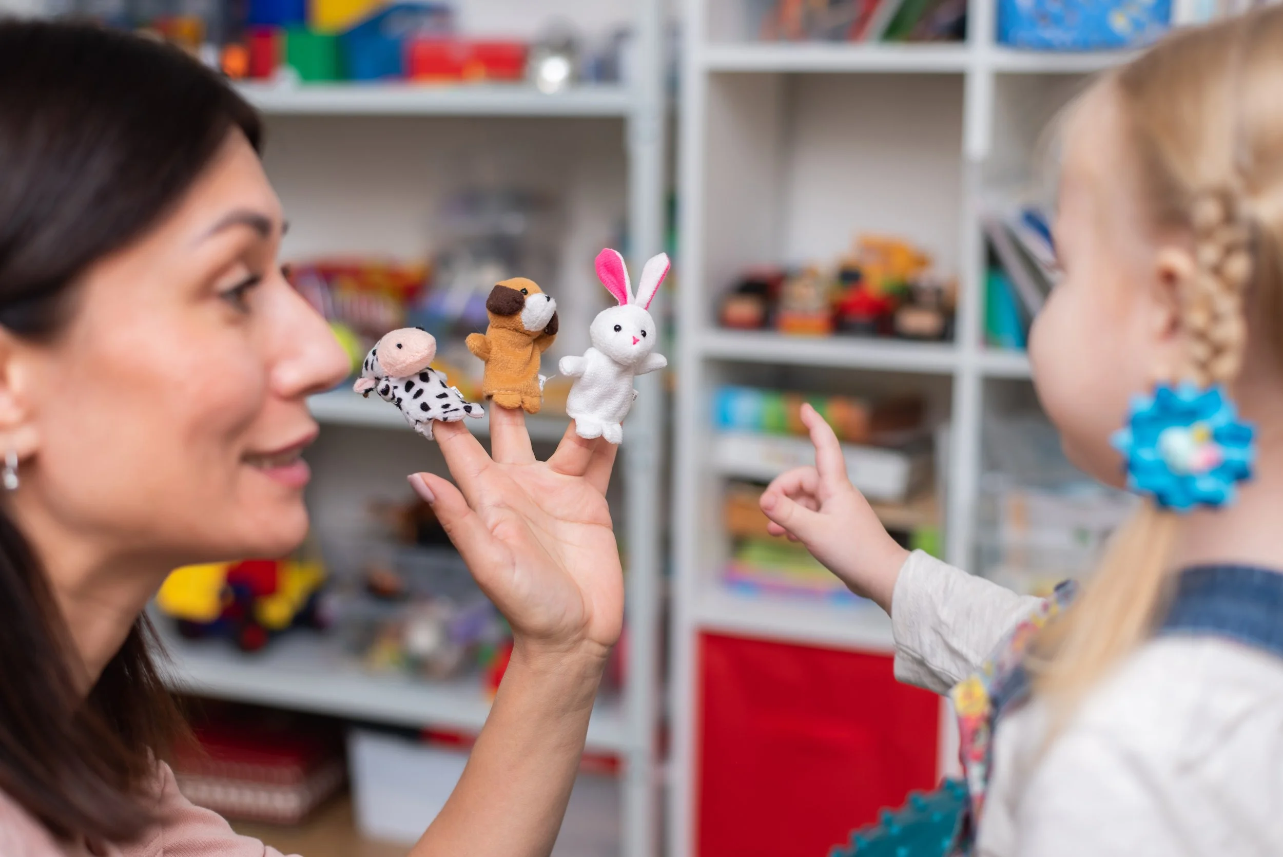 A woman and a girl playing with felt animal finger puppets in a room with shelves of arts and crafts supplies.