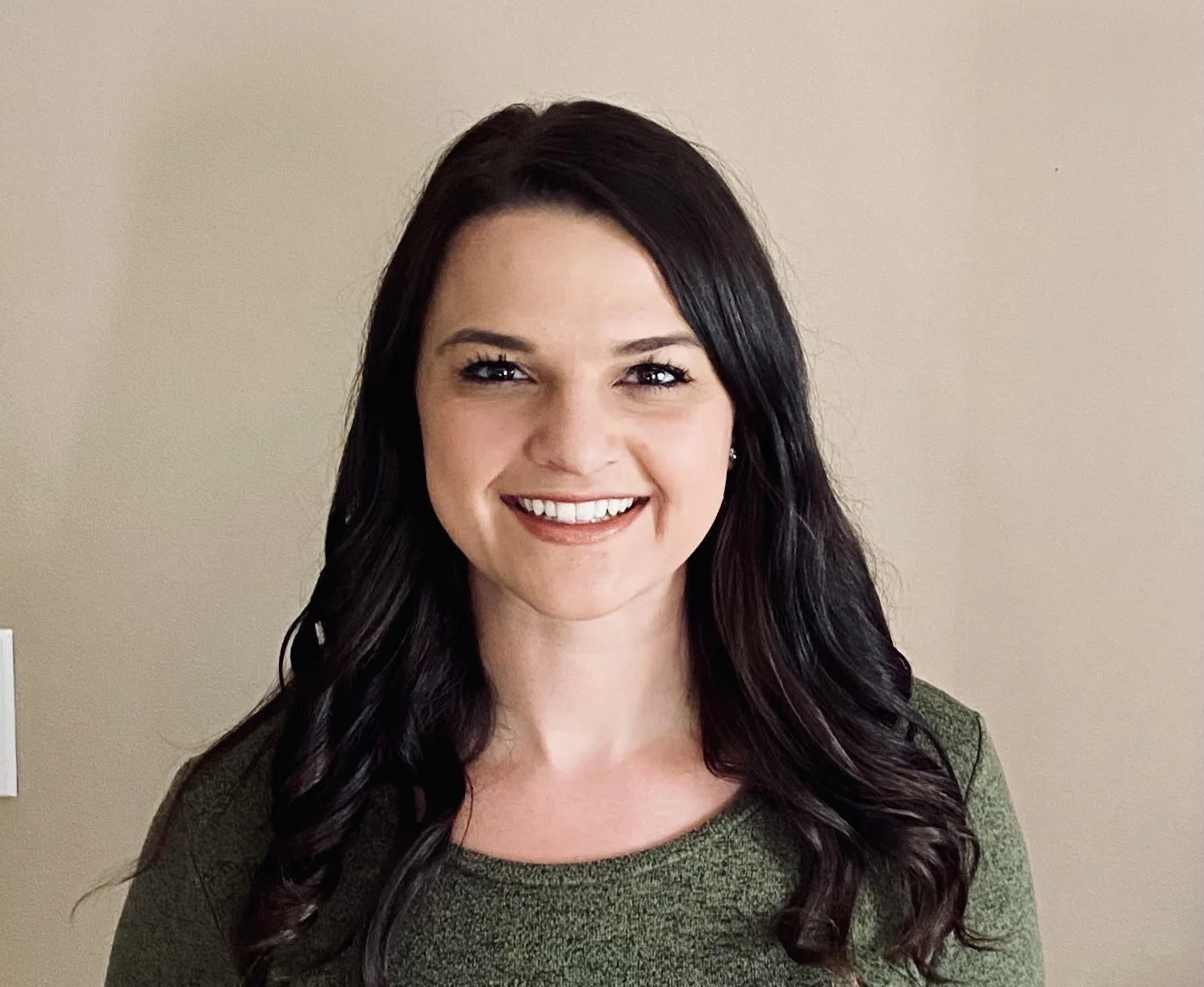A woman with long dark hair smiling, wearing a green top, standing against a beige wall.