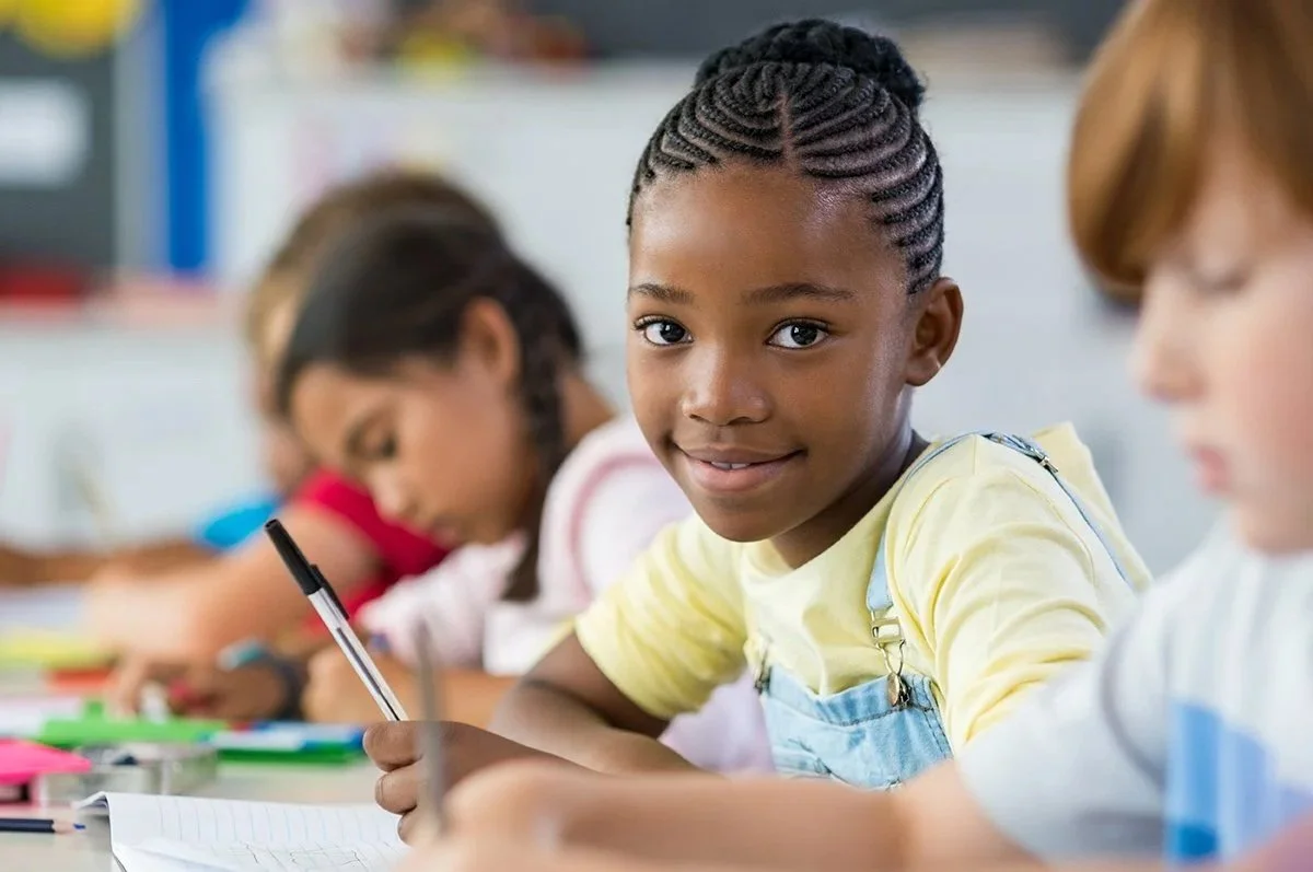 A young girl with braided hair sitting at a desk in a classroom, smiling at the camera, with improved reading and written language skills because of speech therapy for kids in Houston, TX.