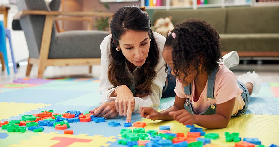 A pediatric speech therapist and a young girl lying on a colorful foam mat, playing with alphabet and number puzzles during in-home speech therapy for kids in Houston, TX.