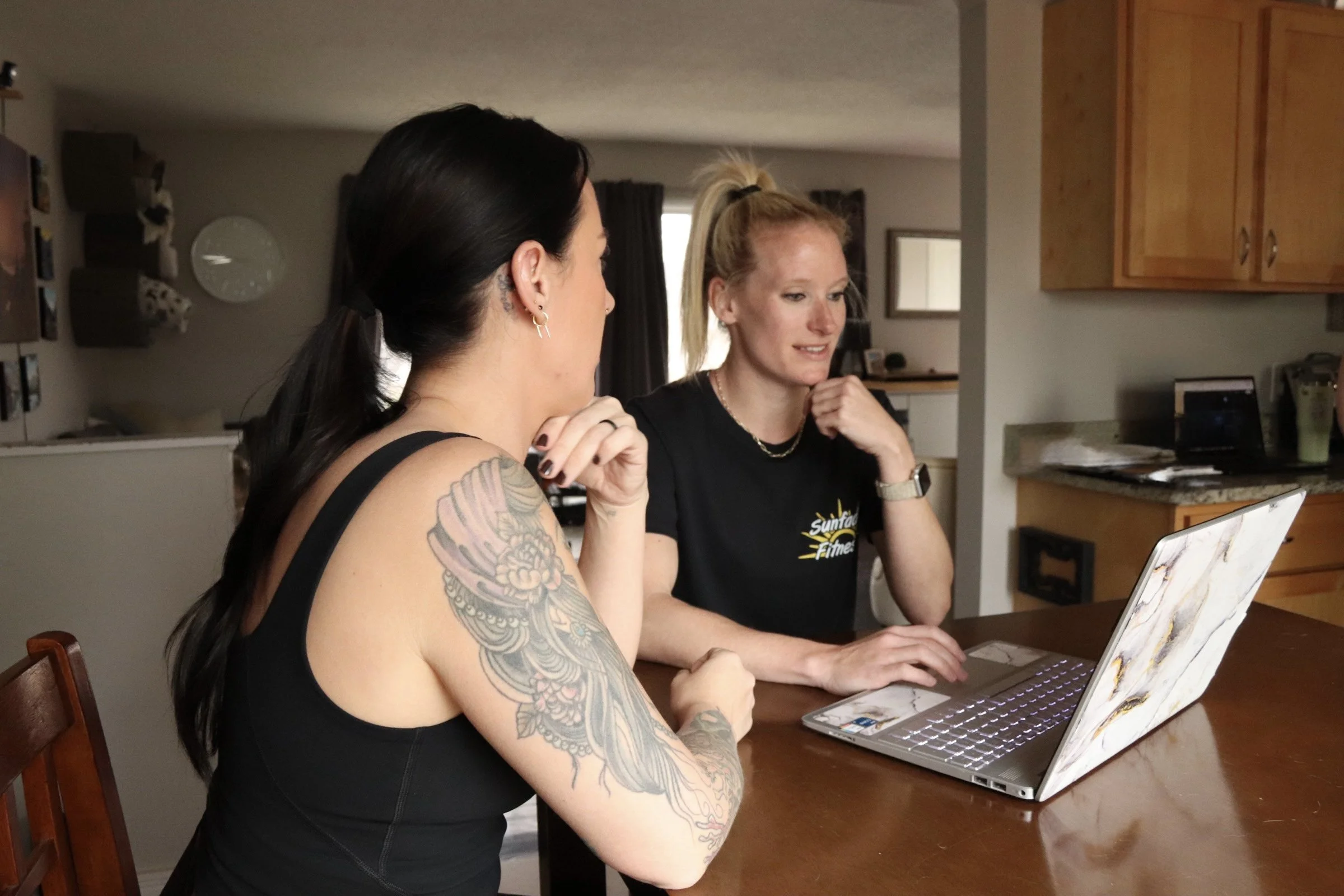Two women sitting at a kitchen table, looking at a laptop, engaged in conversation. One has dark hair in a ponytail and tattoos on her arm, the other has blonde hair in a ponytail, wearing a black T-shirt.