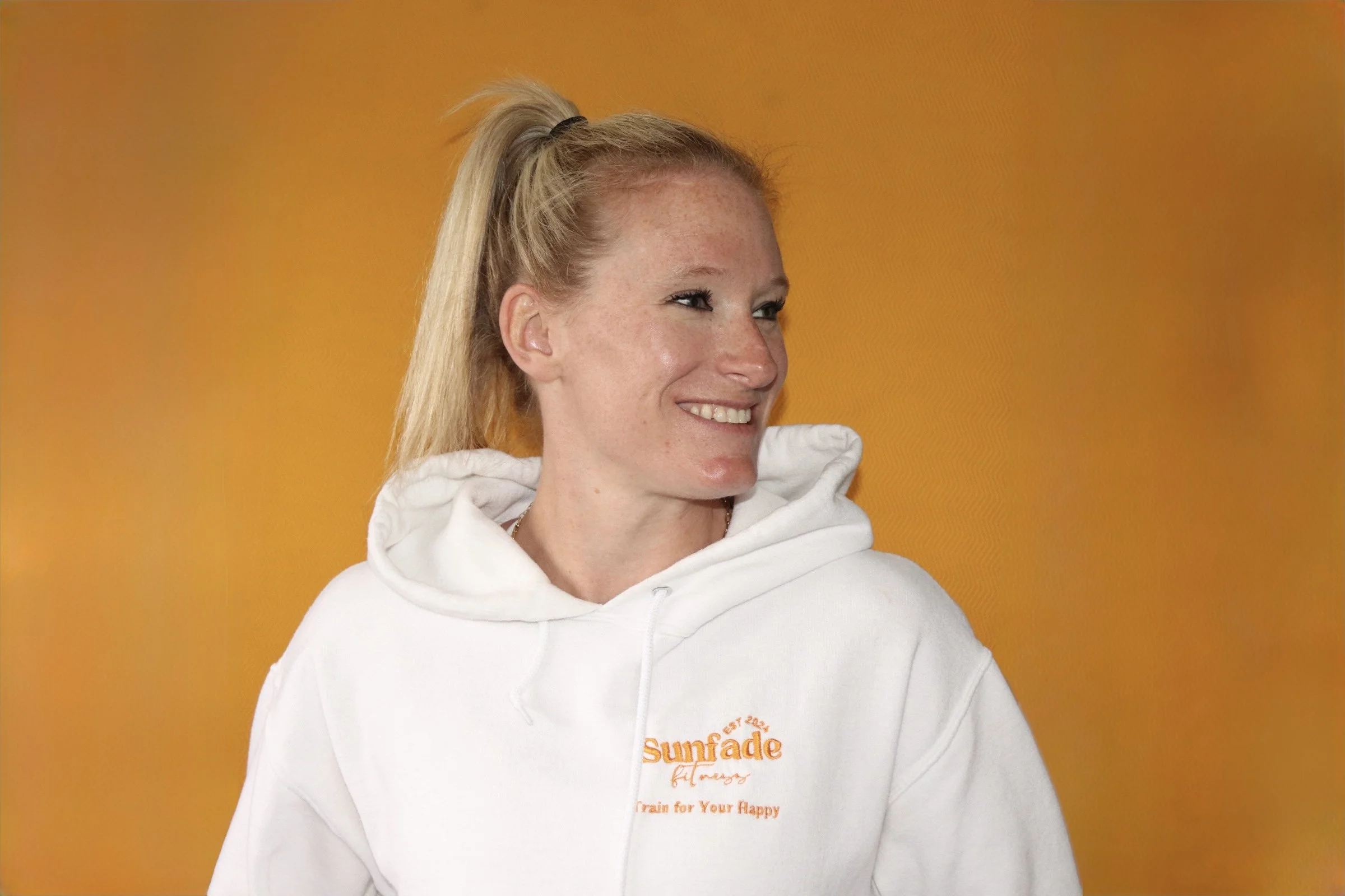 A woman with blonde hair tied in a high ponytail, wearing a white hoodie with yellow text, smiling and looking to the side, standing against a yellow background.