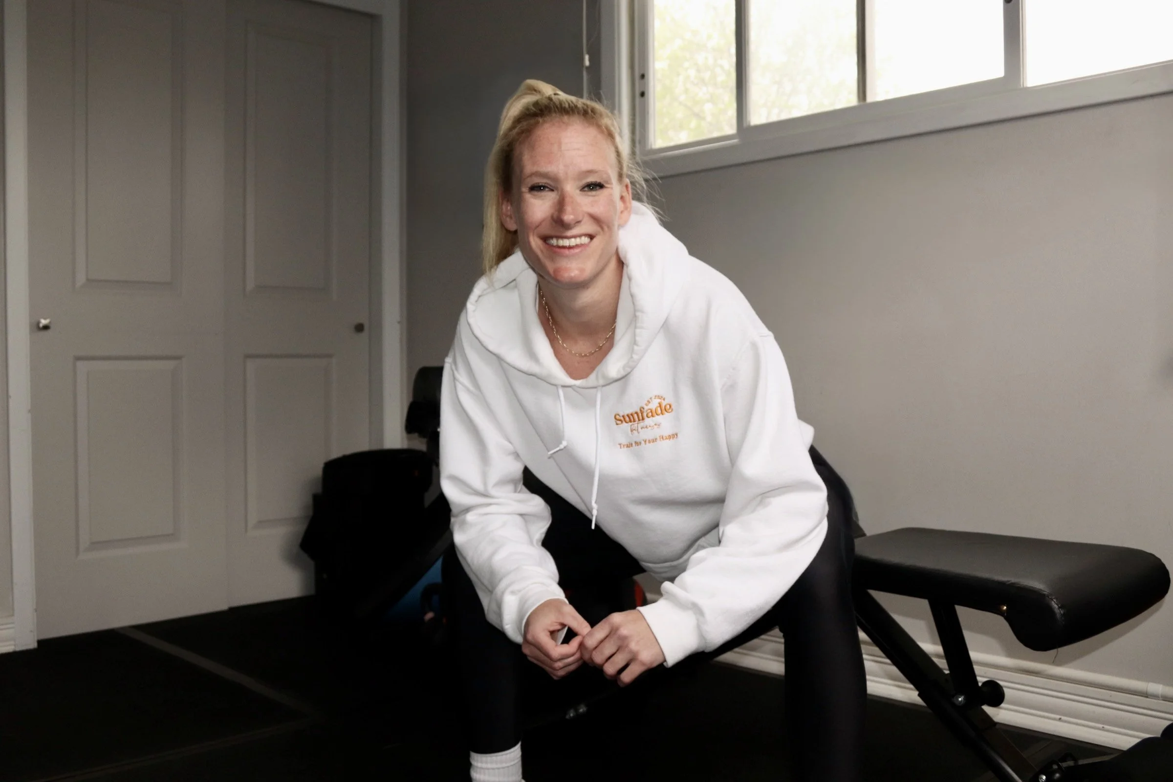 A woman with long blonde hair tied back, smiling, wearing a white hoodie with yellow writing, leaning forward with her hands on her knees, in a room with a window, gray walls, and black chairs.